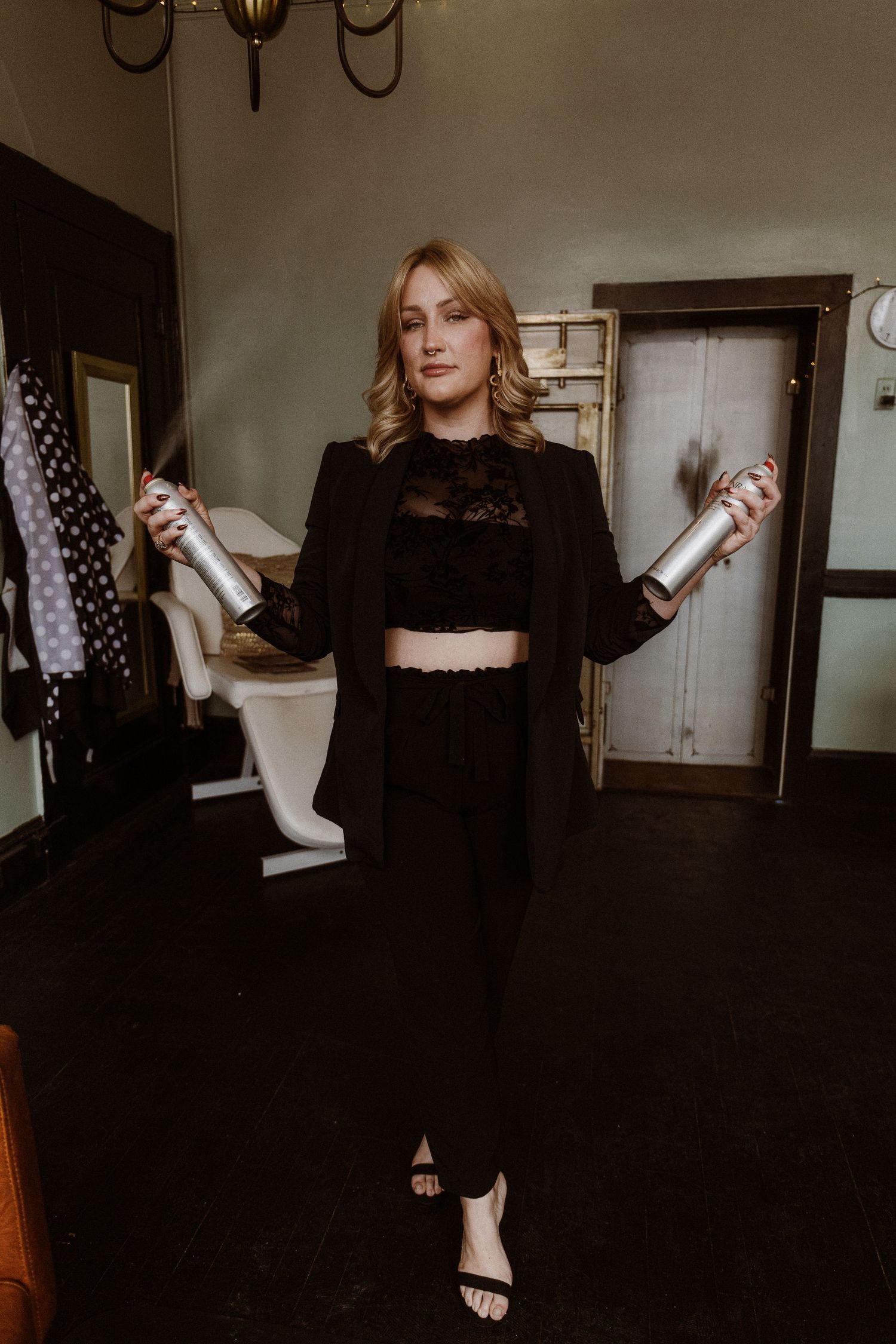 A stylish figure in all black attire poses with bottles against an indoor backdrop.