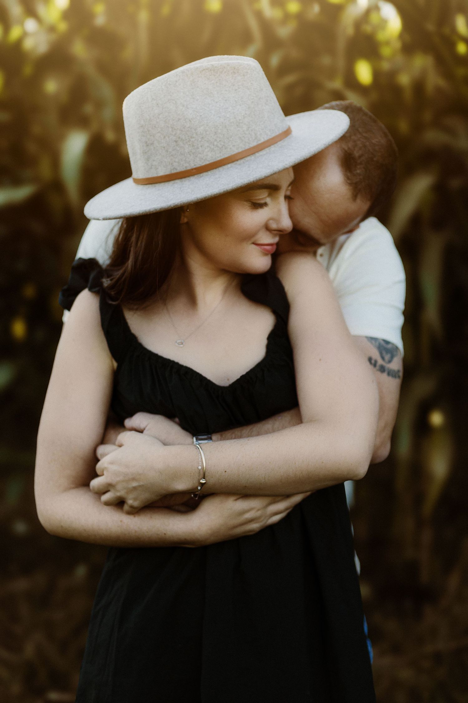 A tender embrace captured outdoors with a white sunhat and black dress in golden light.
