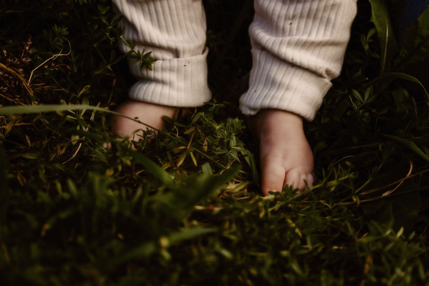Small bare feet in white pants standing on grass in natural lighting.