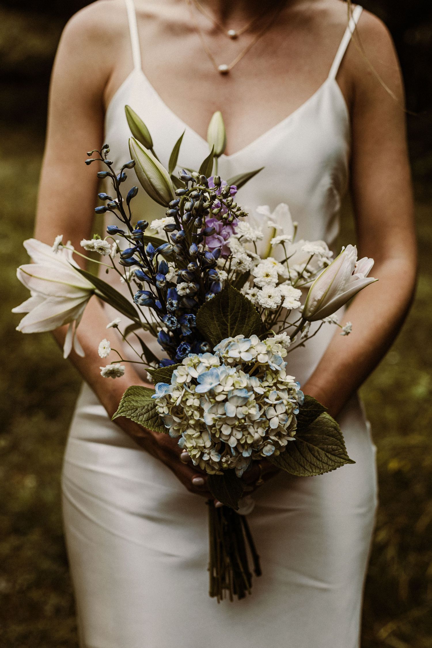An elegant white bridal bouquet with lilies, hydrangeas and blue accents against a white dress.