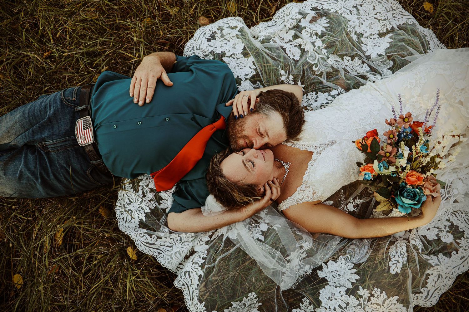 Couple lying on white lace fabric in grass with colorful bouquet.