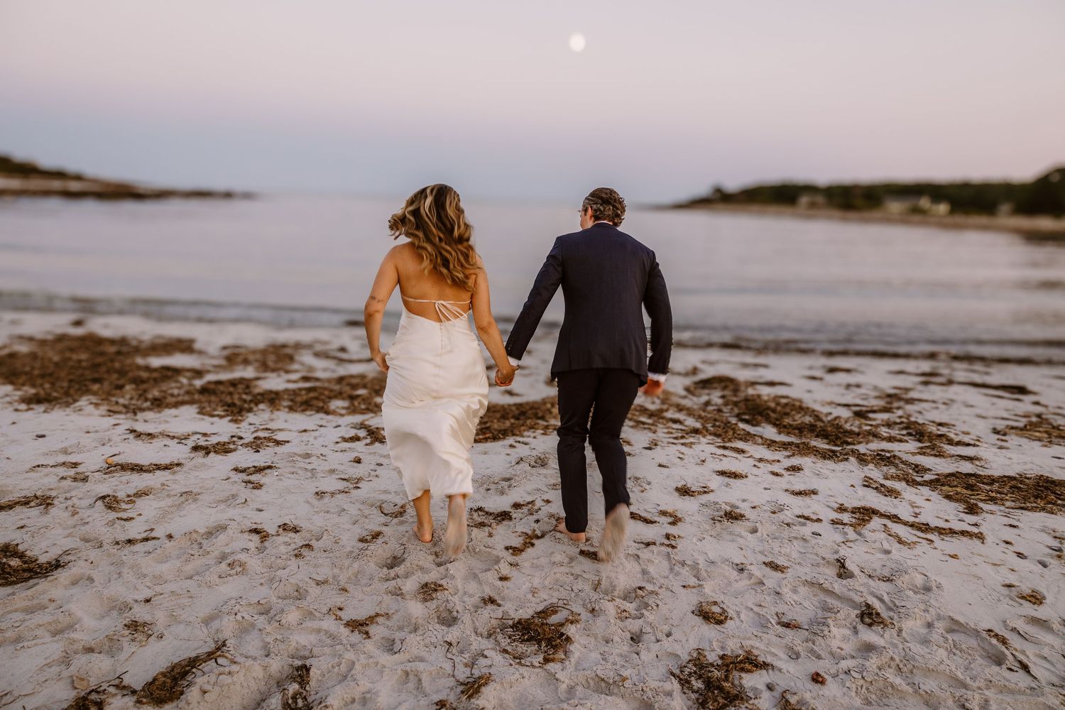 Couple walking hand in hand on a beach at sunset wearing formal attire.