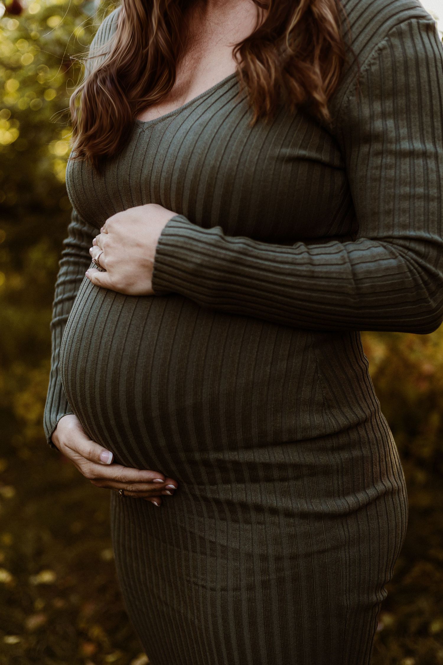Pregnant woman in olive green ribbed sweater dress holds her baby bump against autumn backdrop.