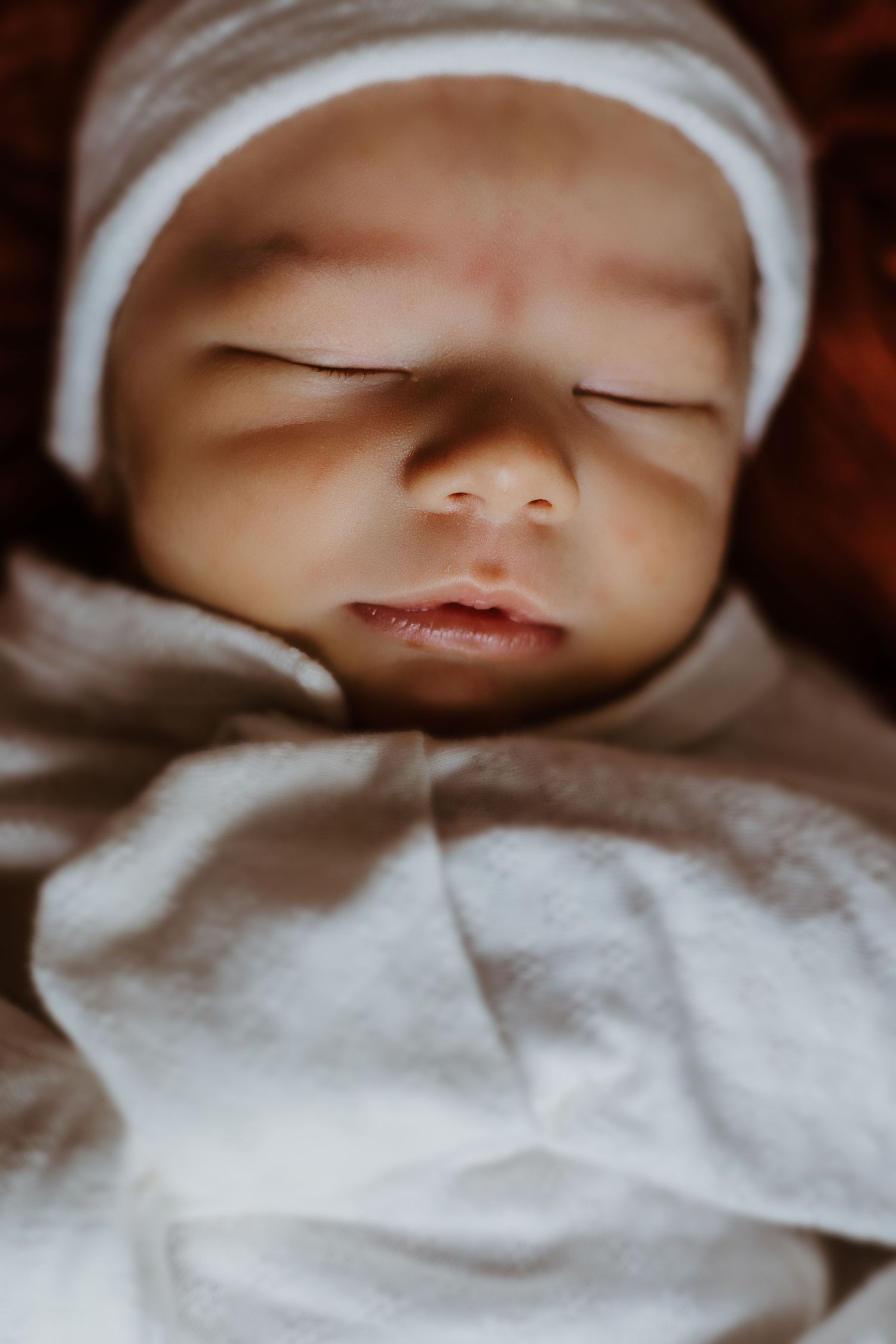 A sleeping newborn baby wearing a white bonnet and swaddled in soft white fabric is photographed in closeup.