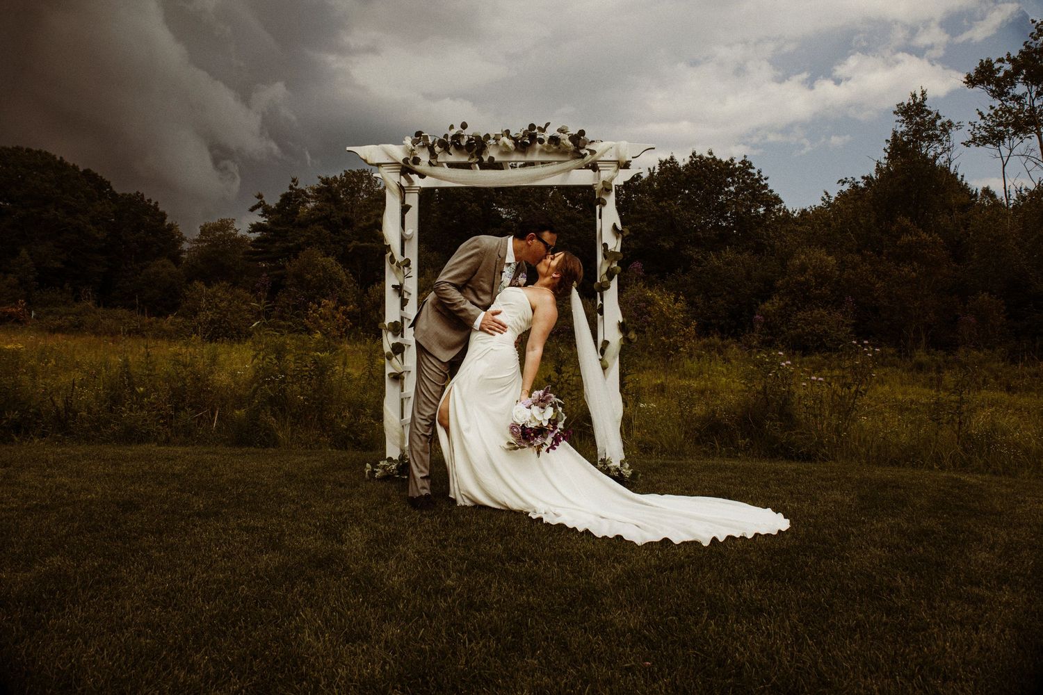 Dramatic wedding portrait under decorated archway with stormy sky background.