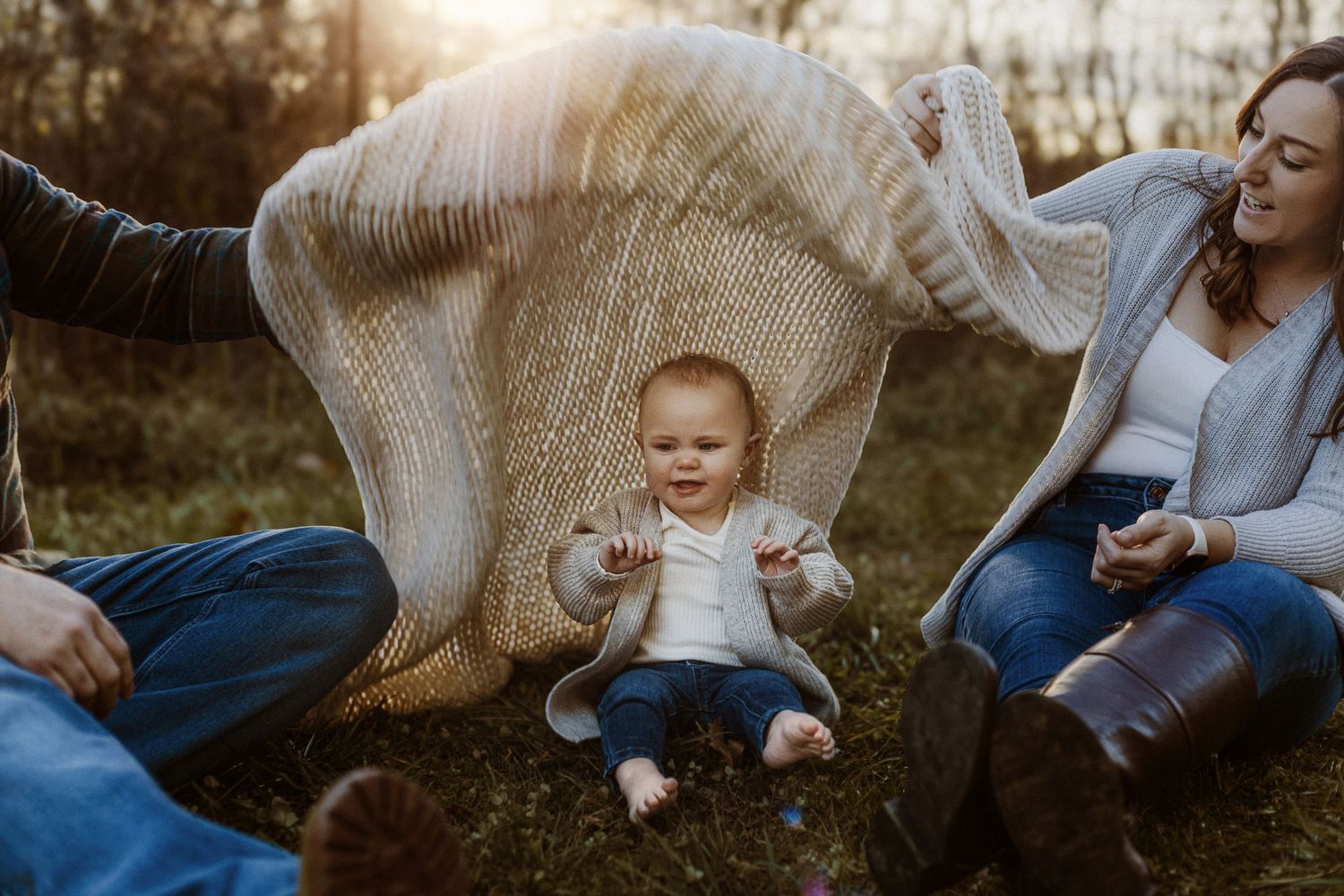 Family throws blanket in air while playing with baby outdoors at sunset.