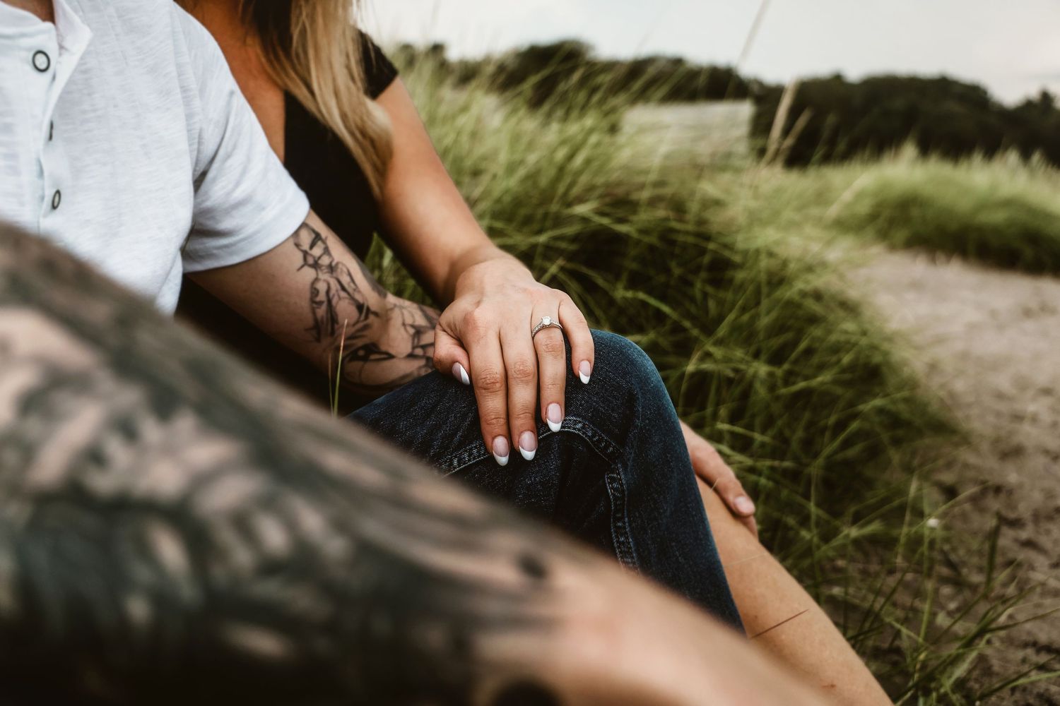 Close up view of tattooed hands resting on denim jeans against a blurred grassy background.