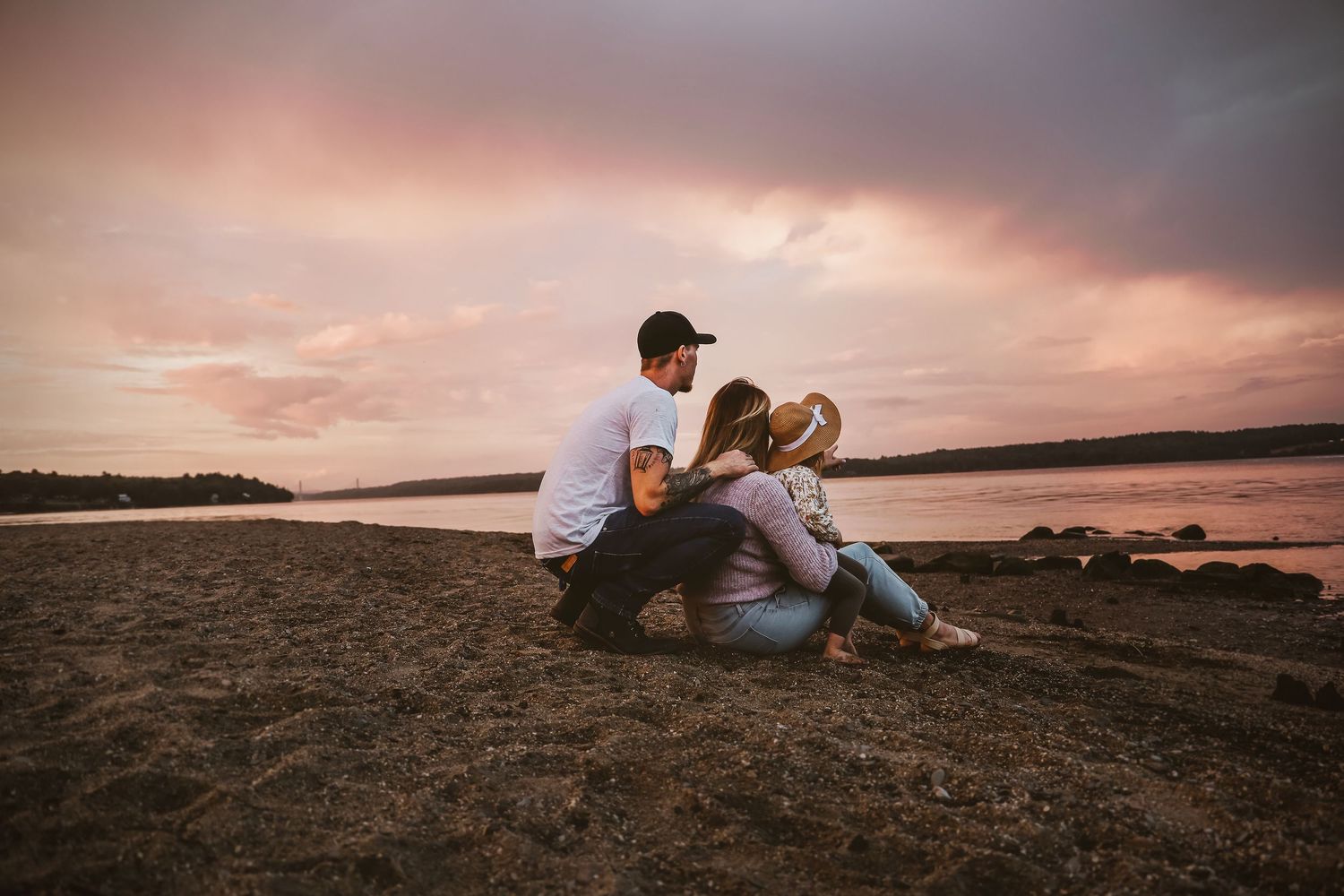 Two people sit together on rocky shore watching a dramatic pink sunset over calm waters.