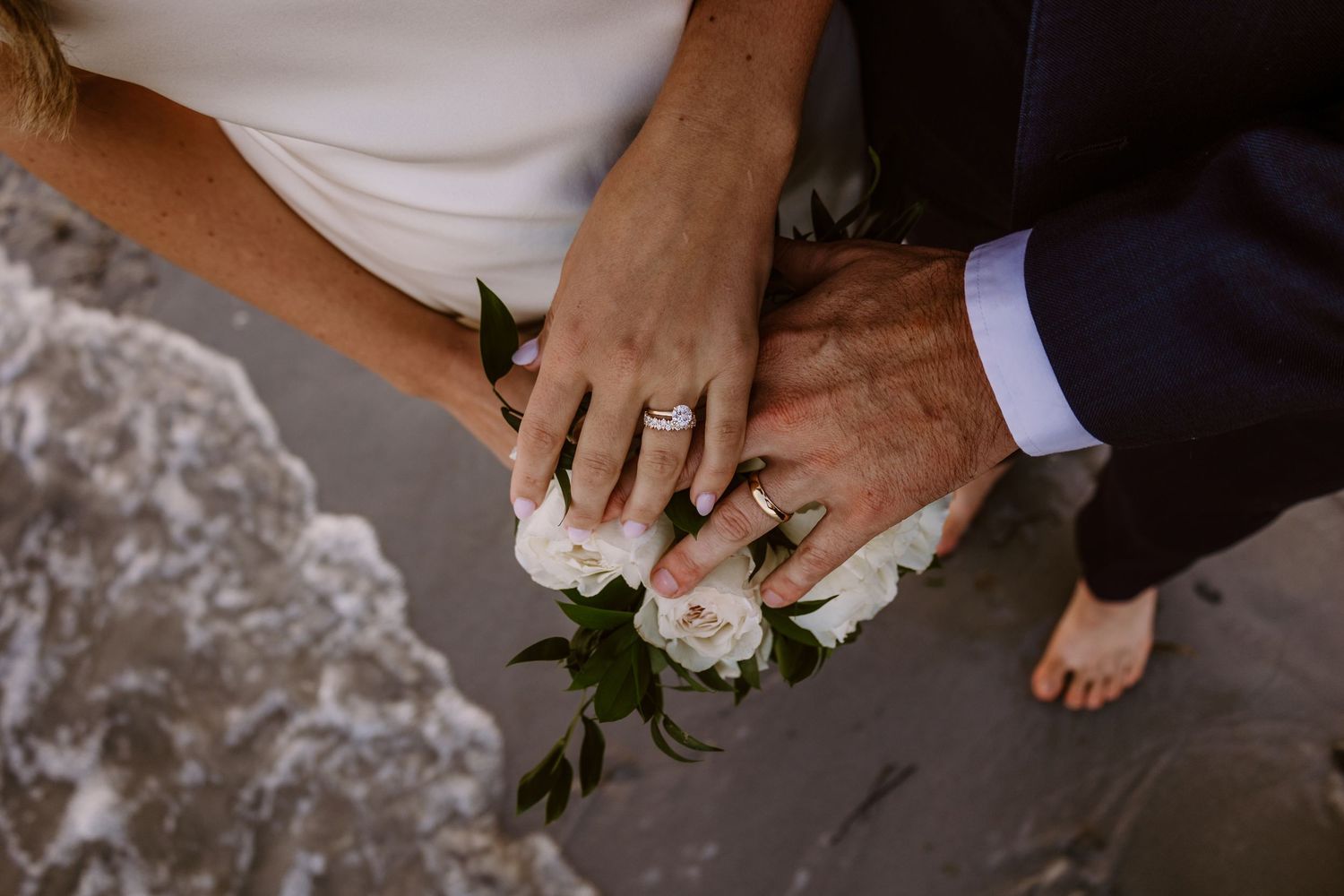 Close-up of wedding rings and hands holding a white rose against sandy beach background.