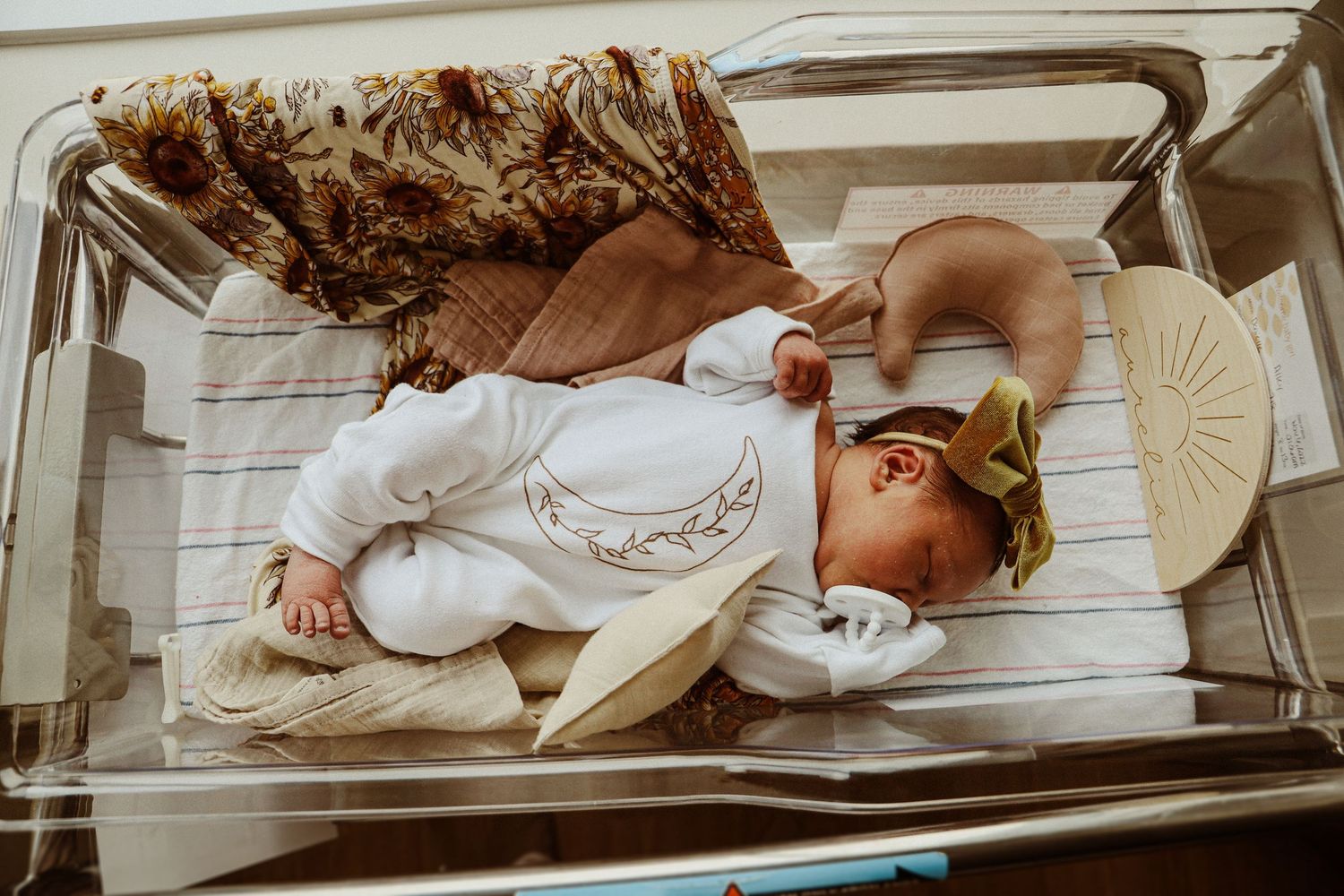 Newborn baby sleeping peacefully in white onesie in hospital bassinet.