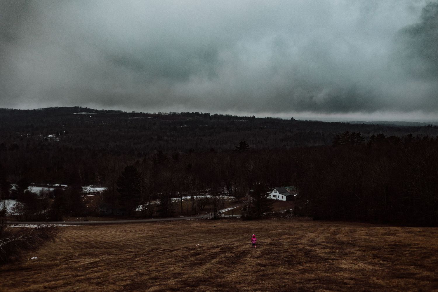 Dark moody landscape with snow patches and distant houses under stormy clouds in rural countryside.