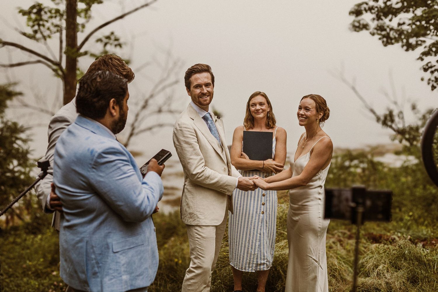 Small wedding ceremony with officiant and guests in natural outdoor setting.