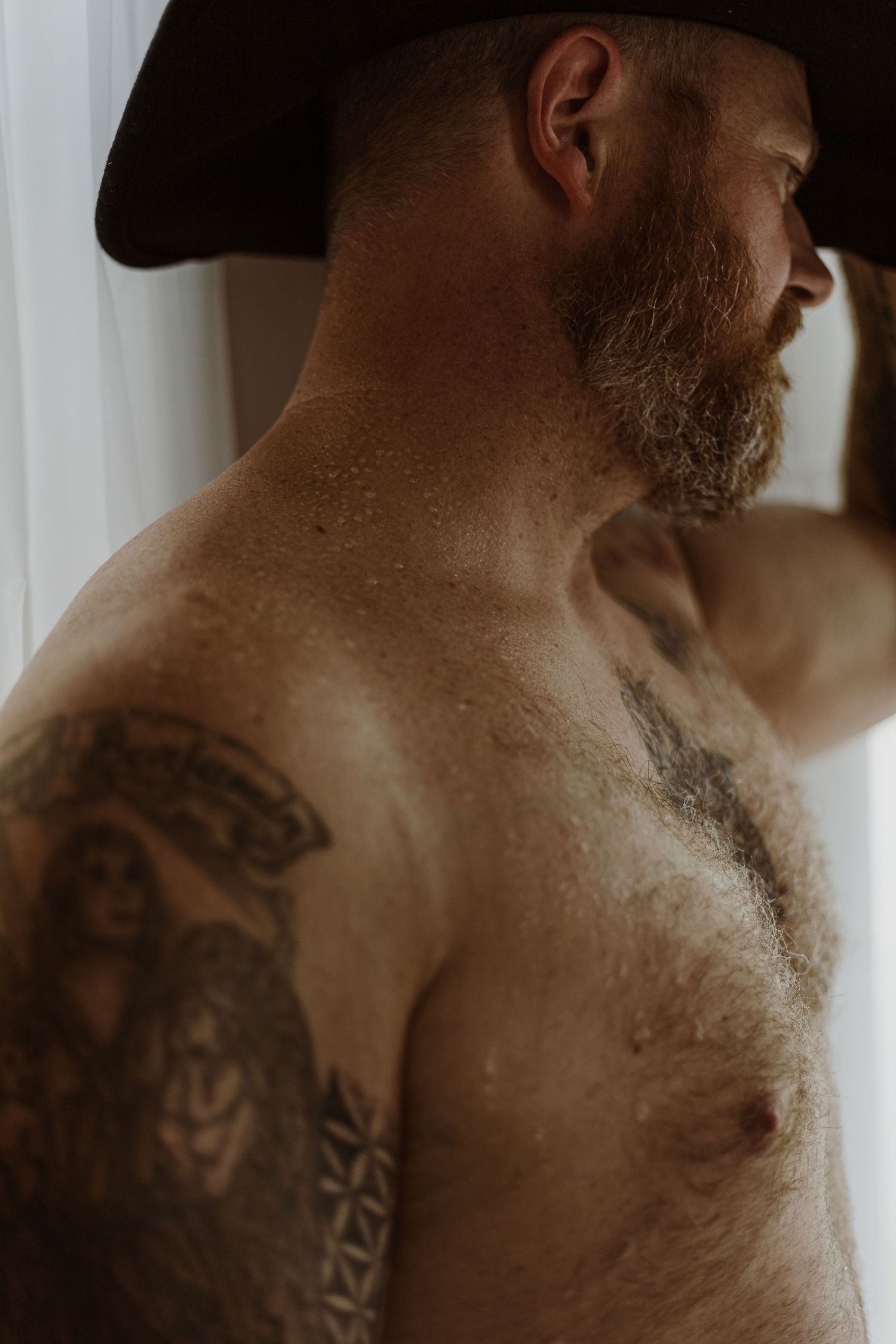 Side view of a shirtless person wearing a hat, showcasing tribal-style tattoos on their muscular shoulder and chest.
