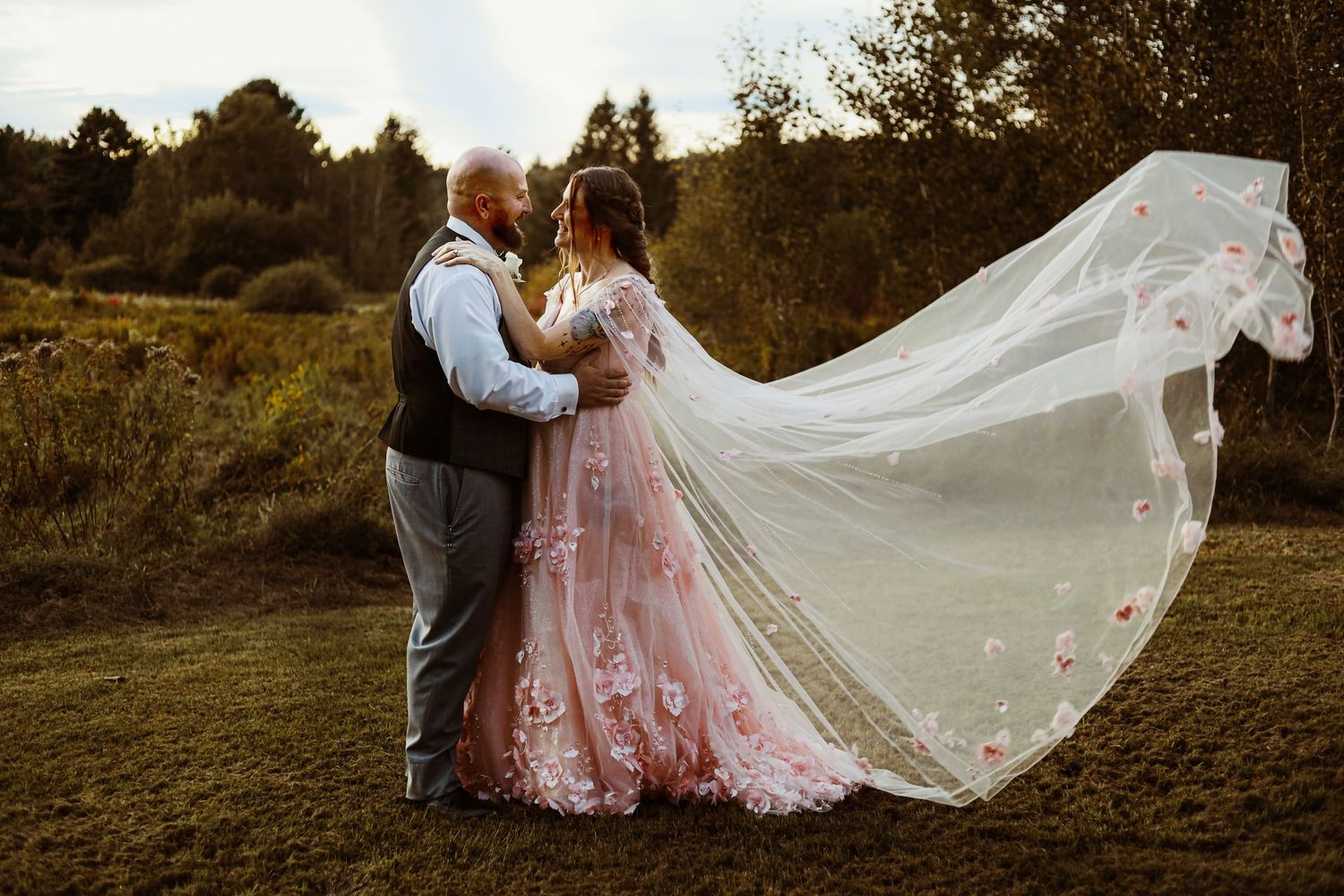 Bride in pink dress with flowing veil poses with groom in sunset field.