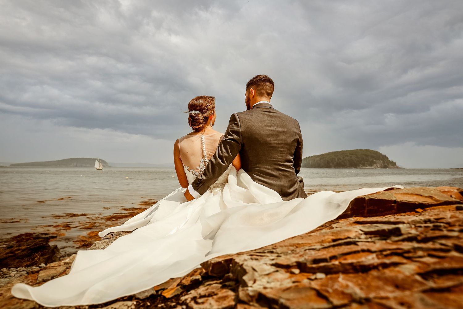 Couple sits together on rocky beach shoreline with flowing white wedding dress.