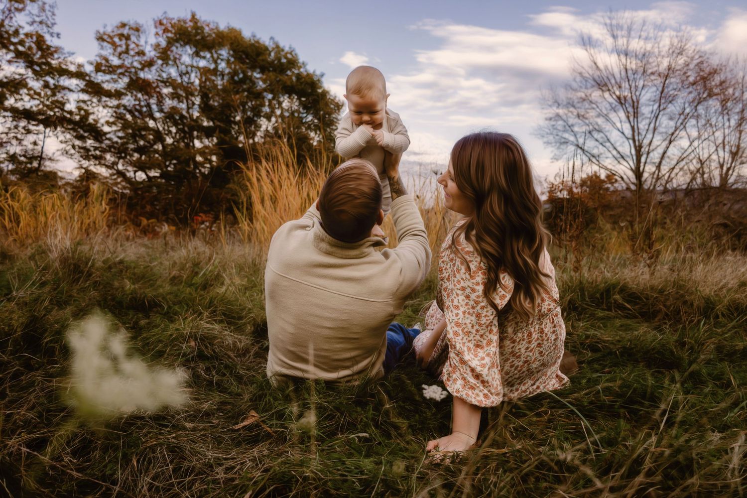 A couple sits in a grassy meadow at sunset with a small child, surrounded by autumn trees and tall grass.