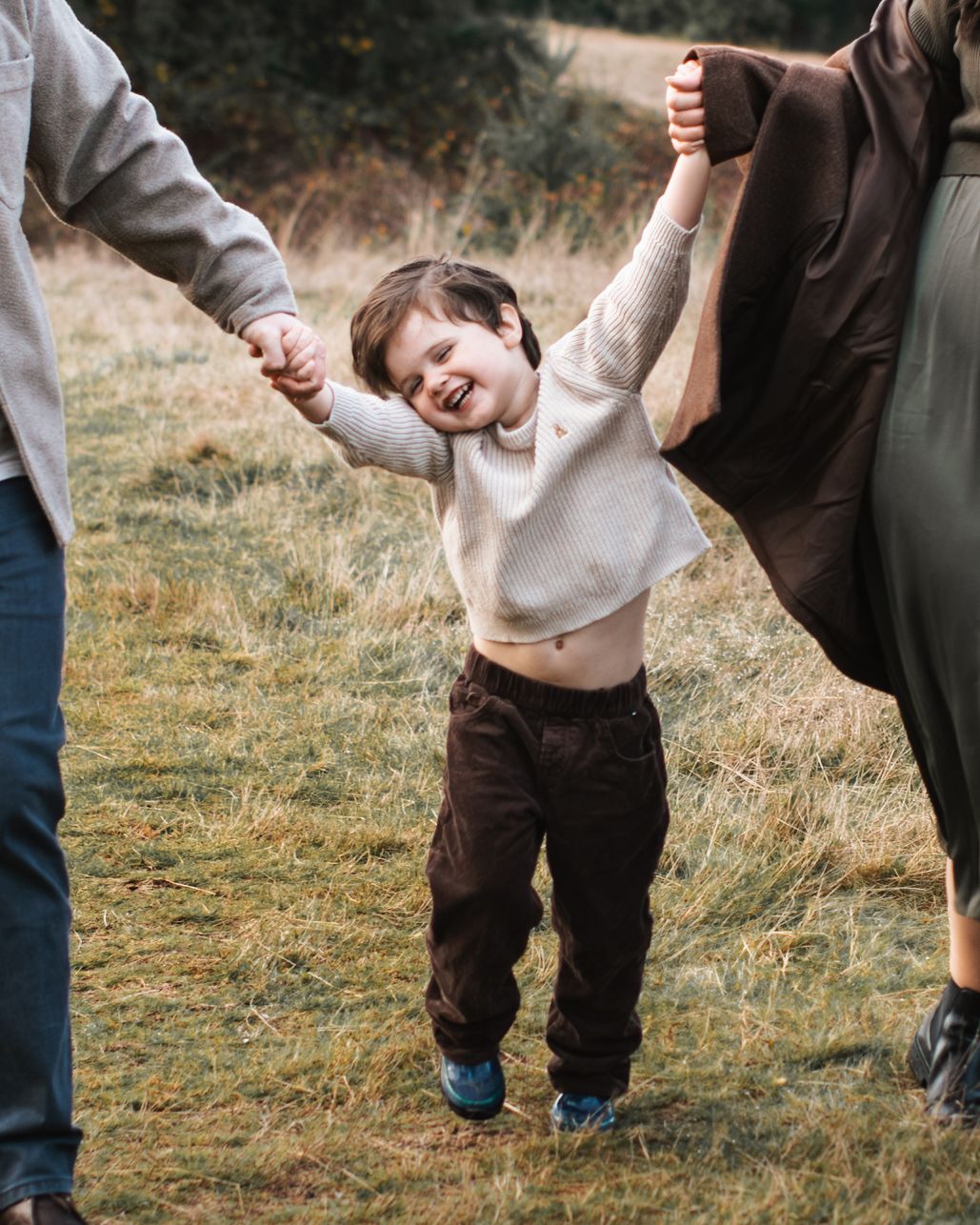 Toddler boy smiling and holding both parents hands during maternity photoshoot and walking in grass