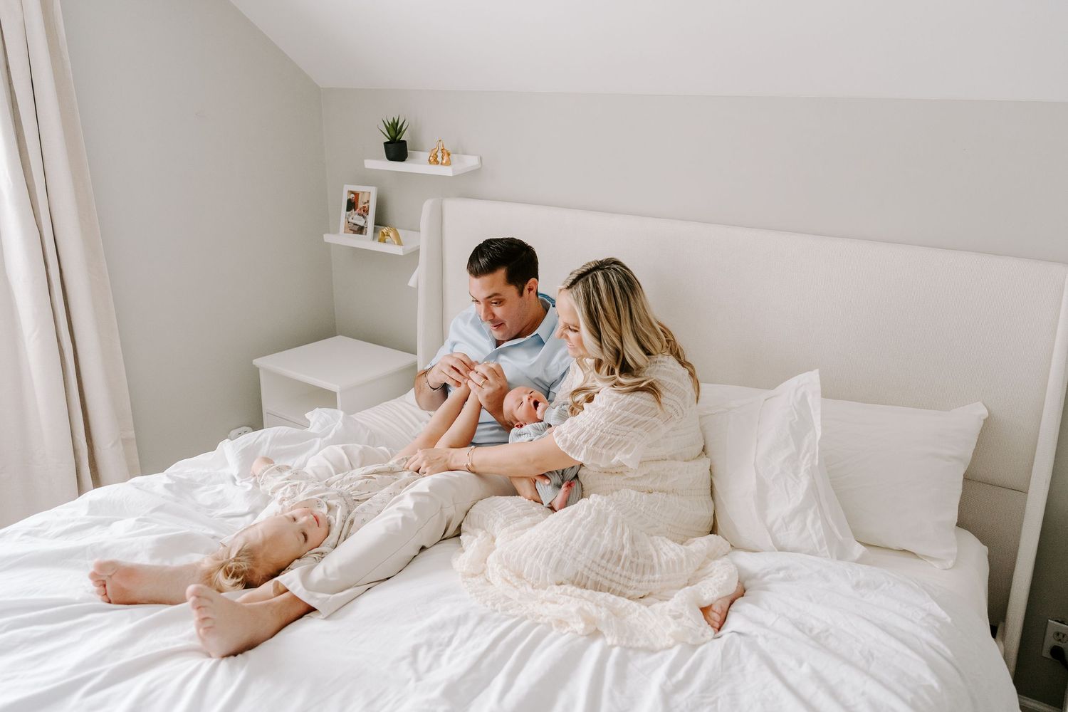 Young couple sitting on white bed holding newborn baby in bright minimal bedroom.