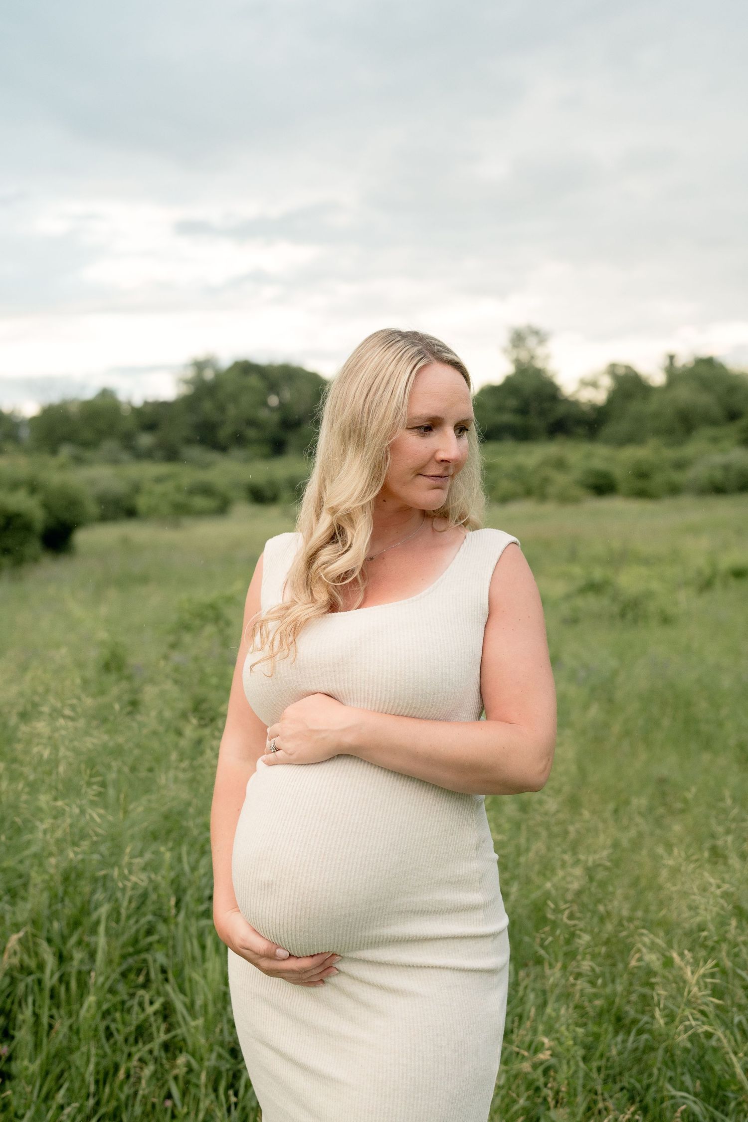 Pregnant woman in white dress cradling baby bump in lush green field, maternity photography outdoors.