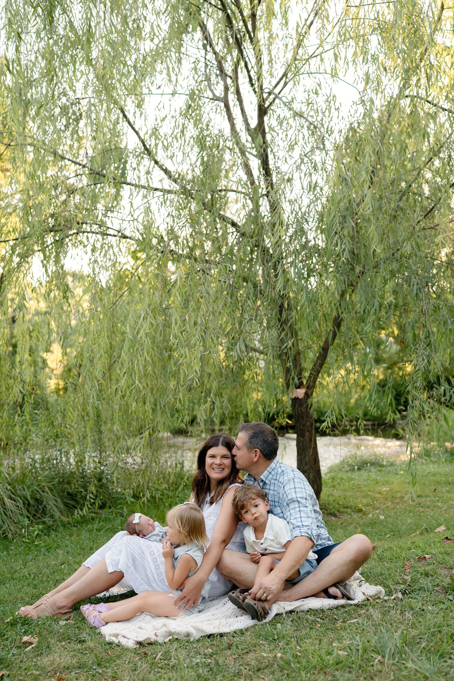 Happy family sitting on blanket under willow tree, parents with three young children enjoying outdoor picnic in green park.