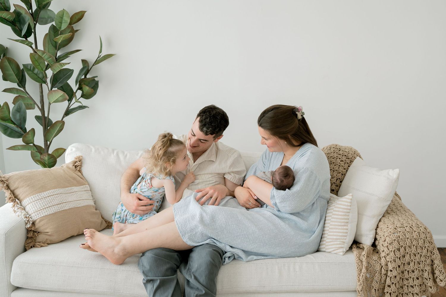 Family of four sitting on a white couch, parents with toddler and newborn baby in a bright living room.