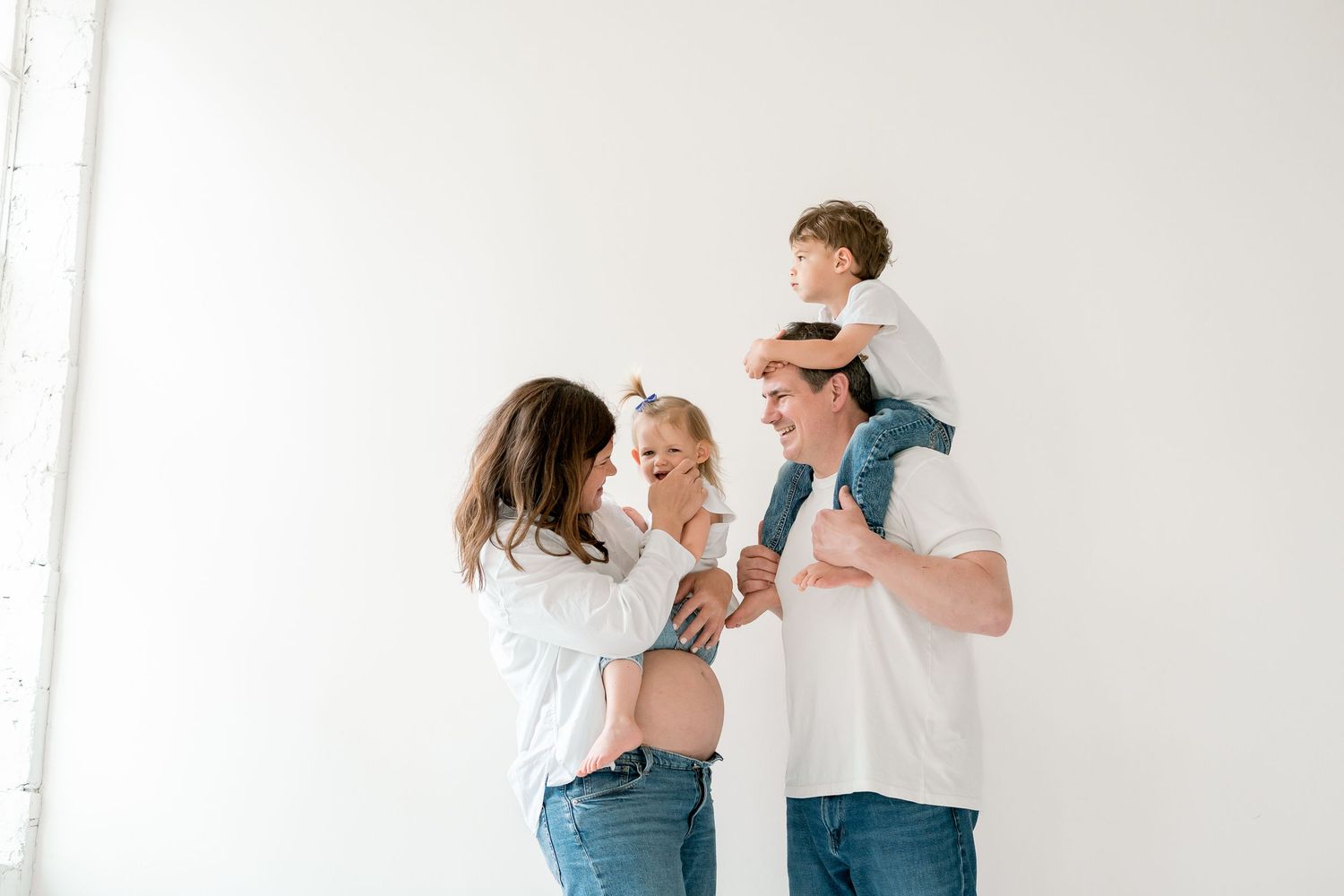 Happy family of four in white shirts and jeans posing together, with toddler on dad's shoulders and pregnant mom holding baby girl.