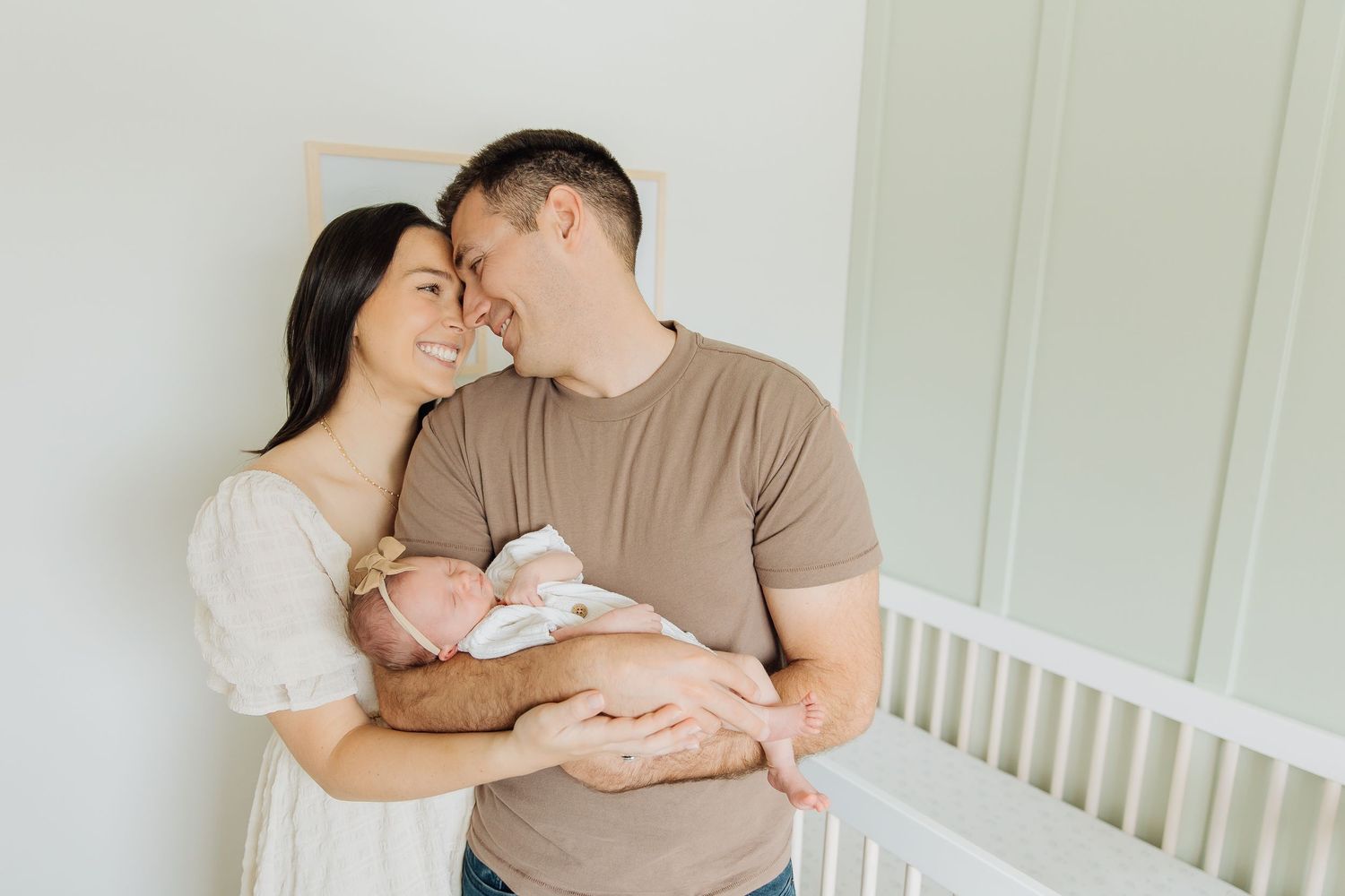 New parents standing together holding their newborn baby in a bright white nursery room.