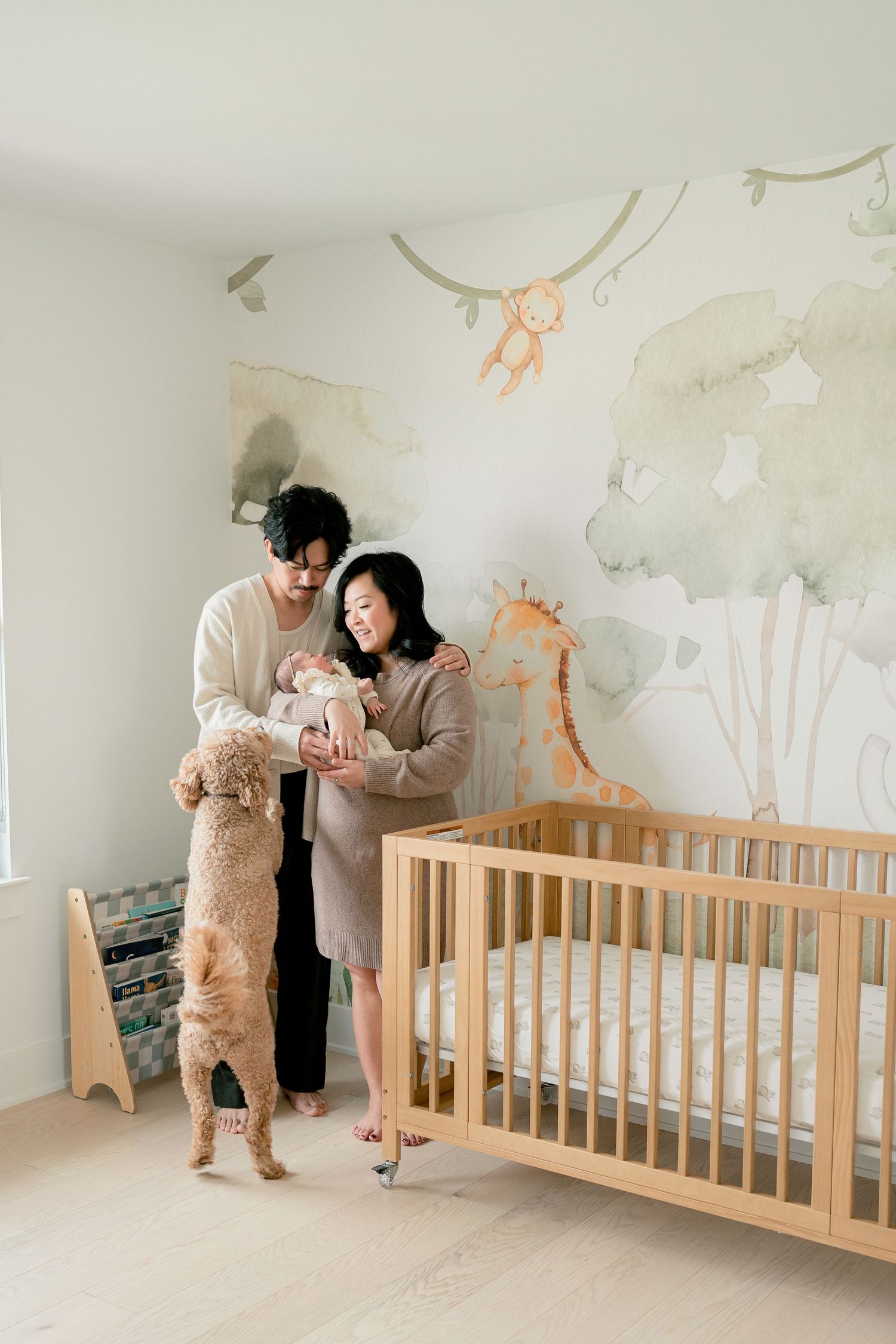 Couple admires newborn baby in safari-themed nursery while golden doodle dog stands on hind legs nearby.