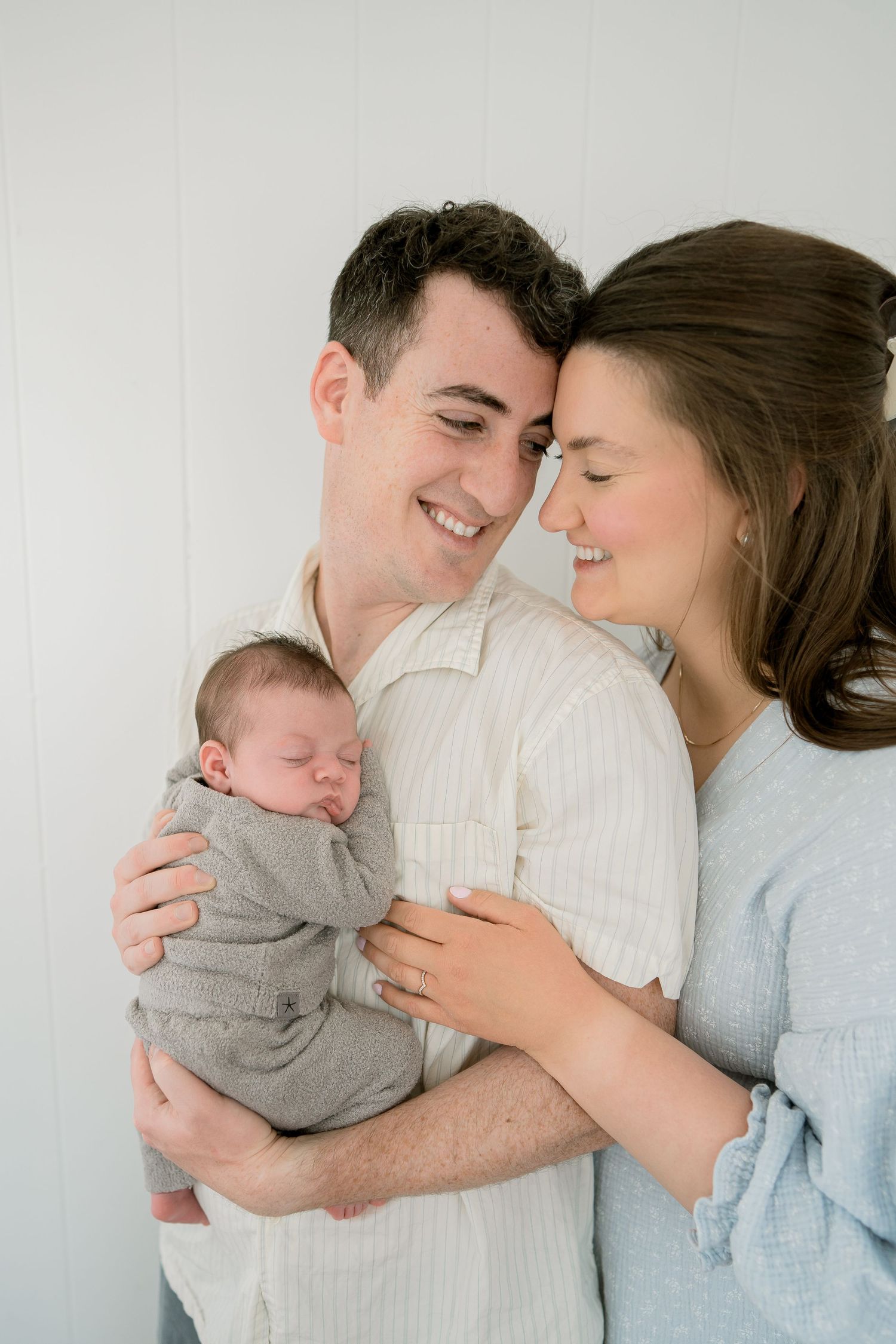 Smiling parents holding their sleeping swaddled newborn baby against a white background.