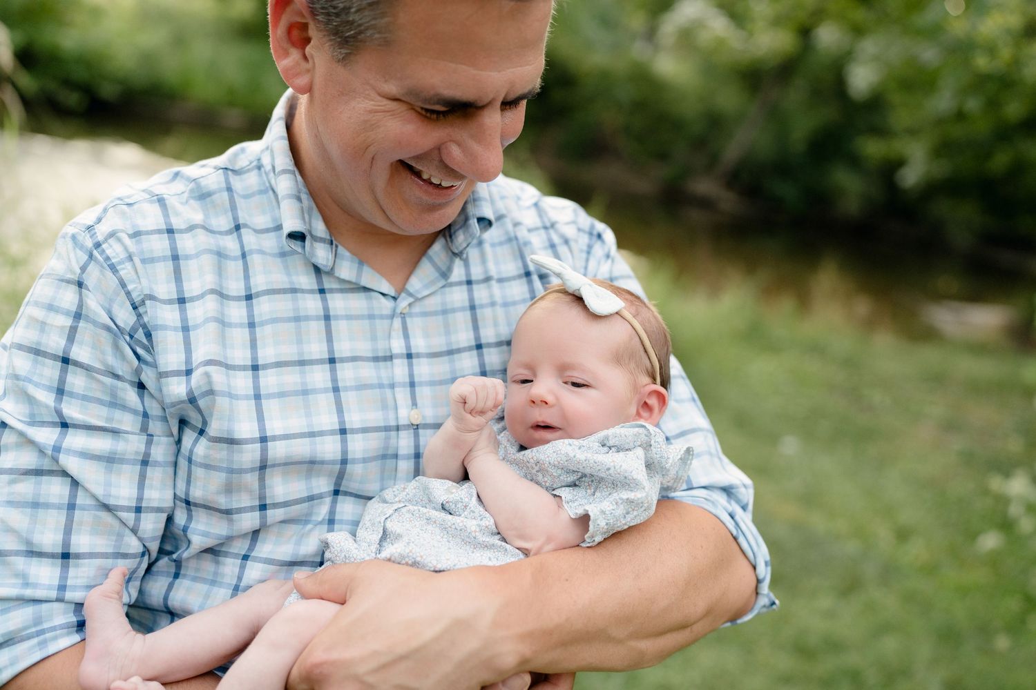 A smiling man in a plaid shirt cradles a newborn baby outdoors in a lush green setting.