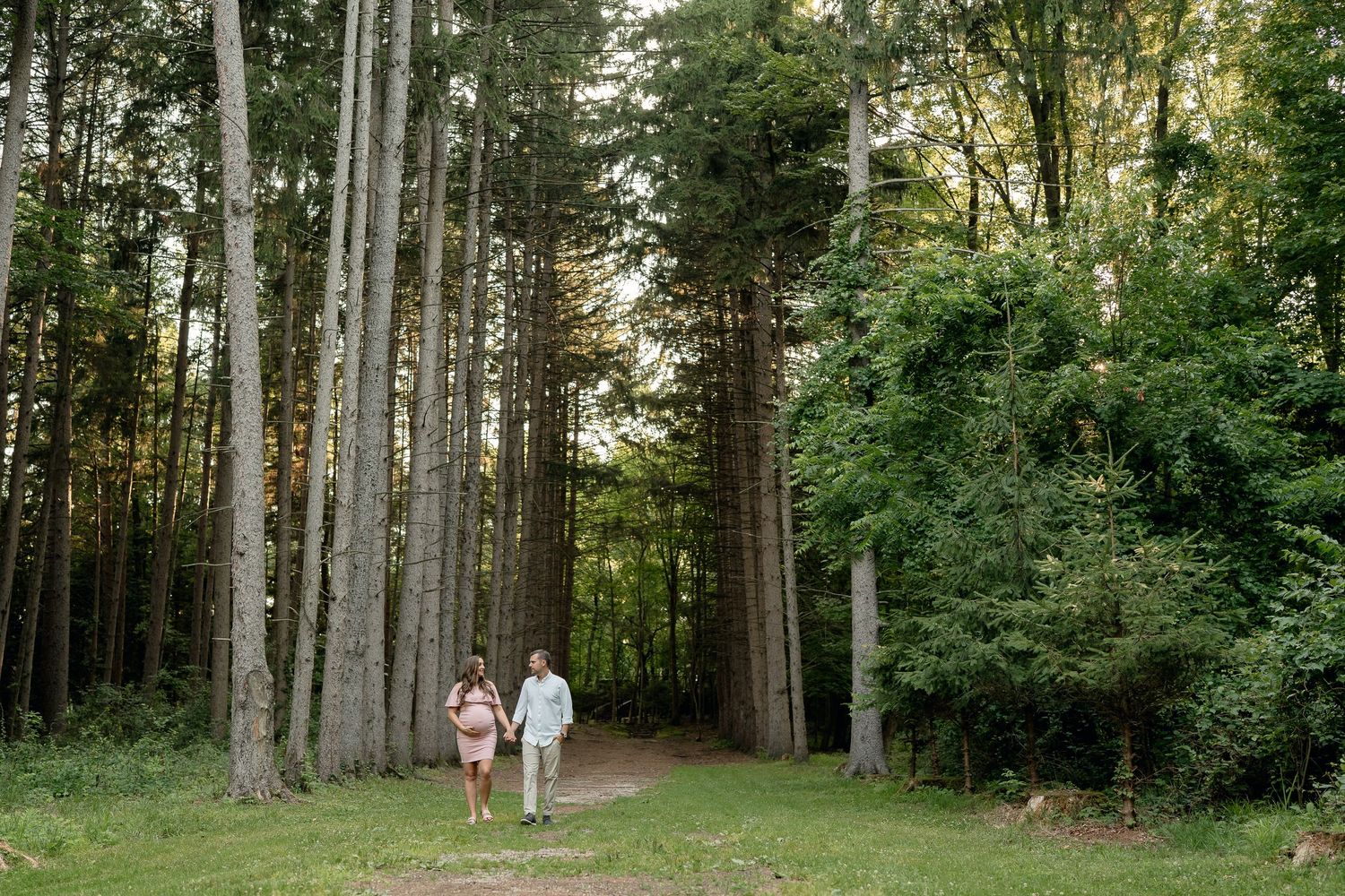 Pregnant couple walking hand in hand along a scenic forest path during an outdoor maternity photography session.