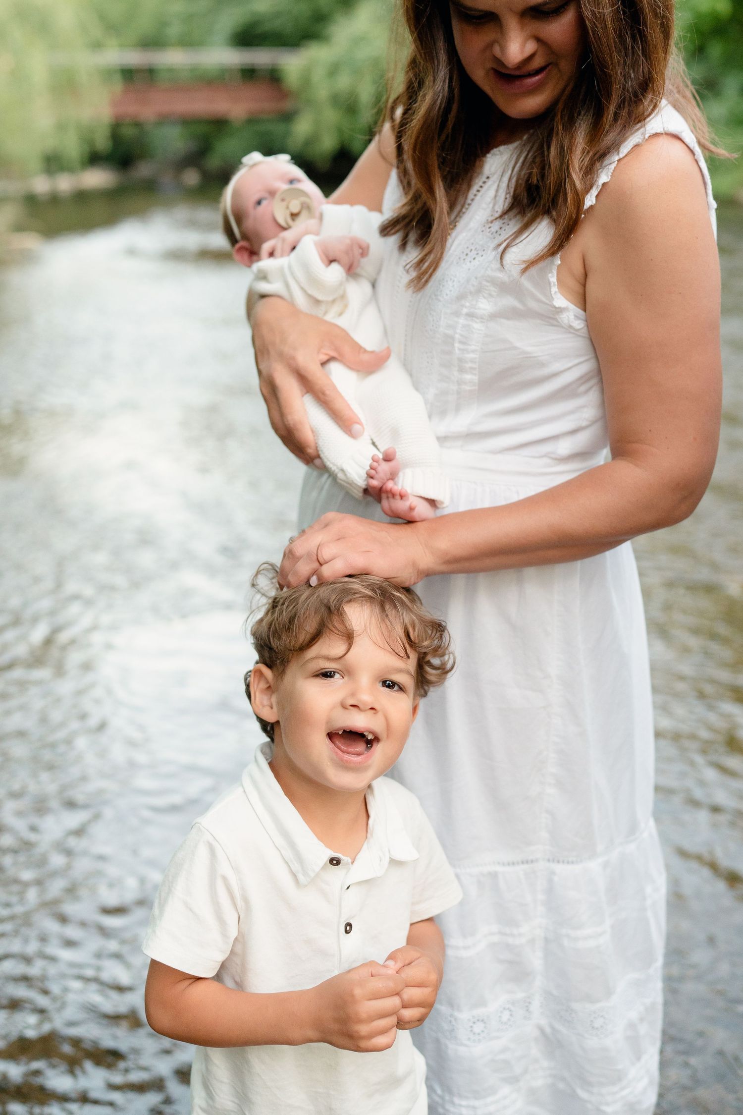 Smiling boy stands in front of mother in white dress holding newborn baby by a creek.
