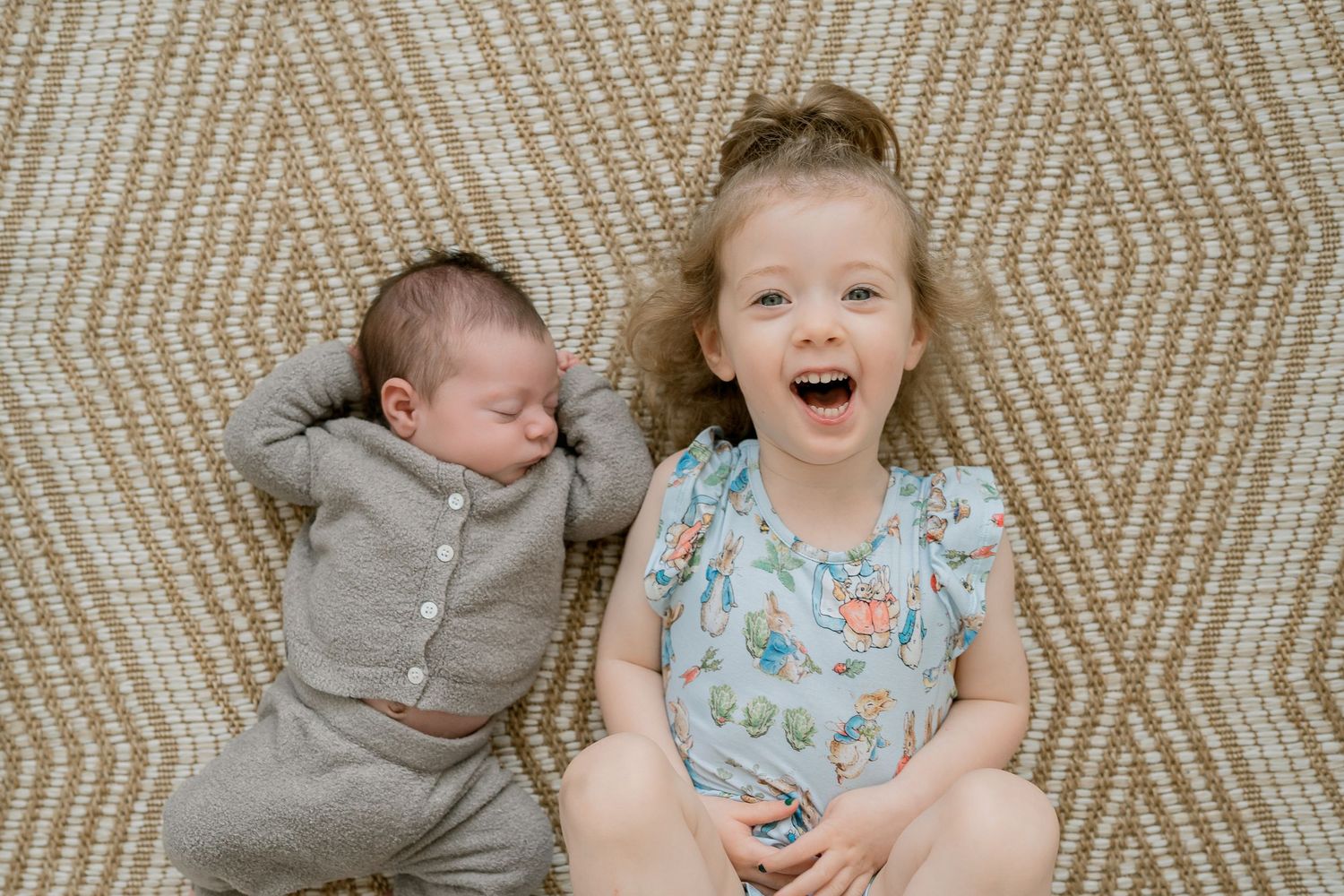 A joyful toddler girl sits beside a sleeping newborn baby on a woven rug, both dressed in casual outfits.