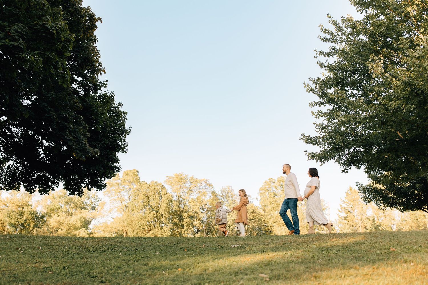 A family runs playfully across a grassy hill framed by large trees on a sunny day.