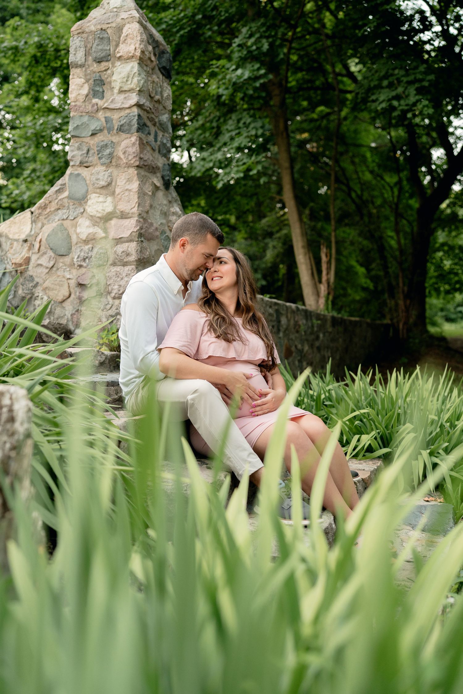 Loving couple sitting together on stone steps surrounded by greenery during a romantic outdoor maternity photo session.