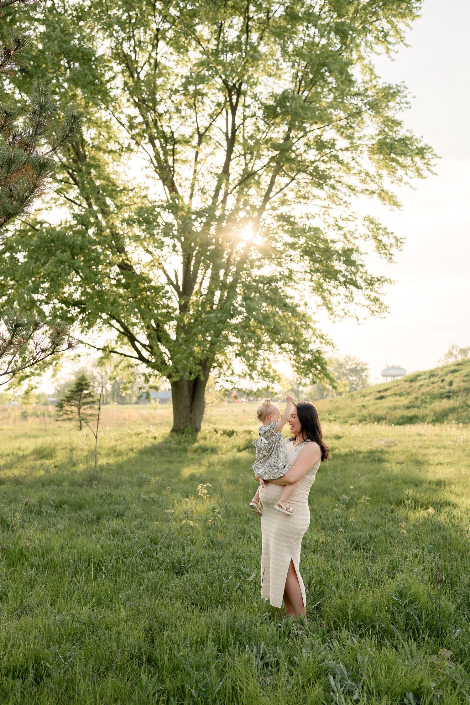 Mother holds smiling toddler under a large sunlit tree in a green meadow.