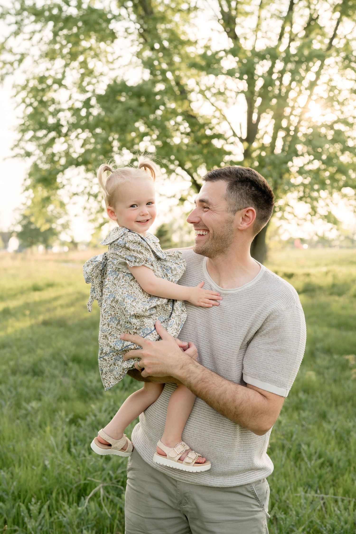Smiling father holding laughing toddler girl in floral dress in sunny green field.