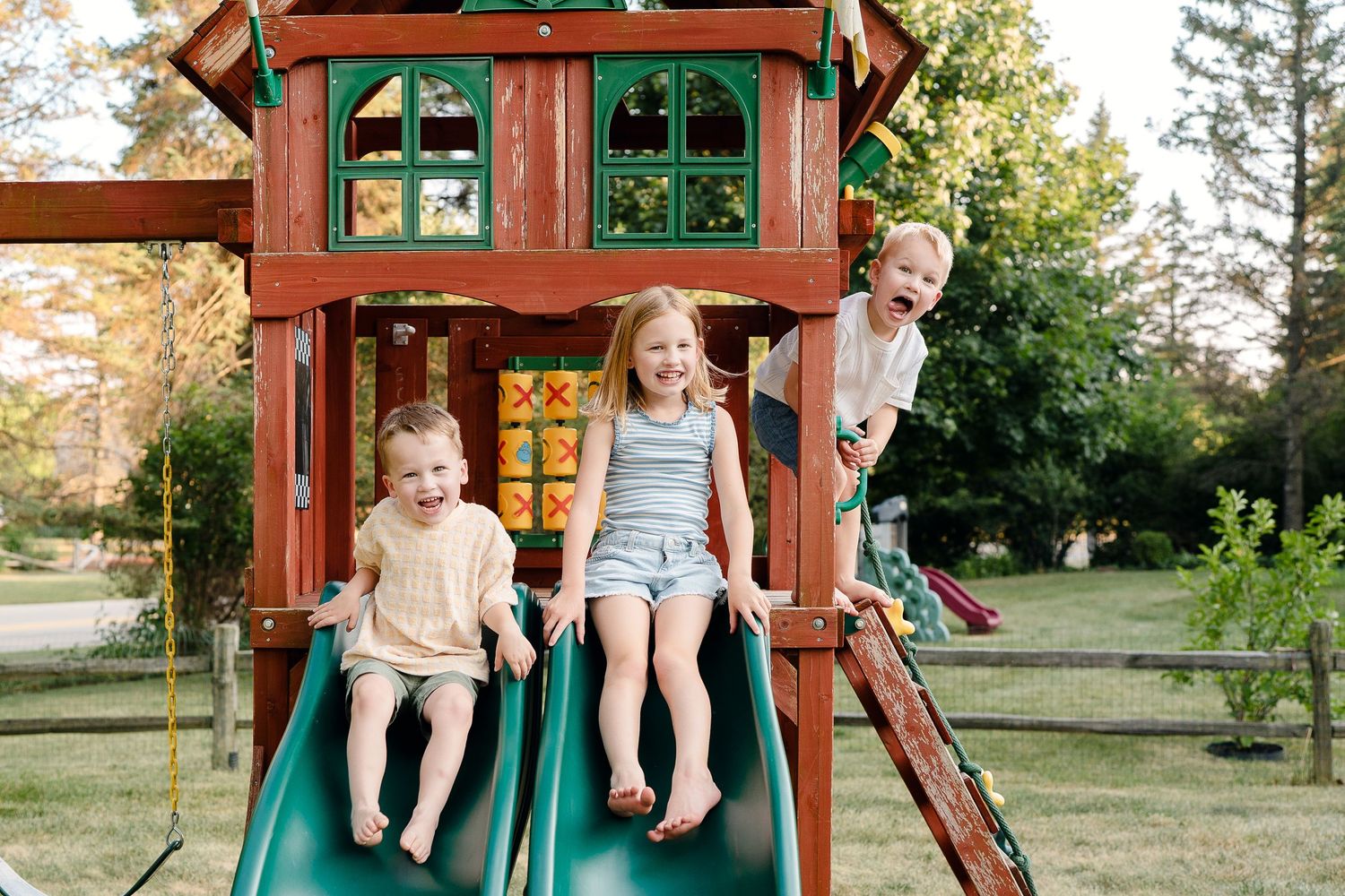 Three happy children sitting on green slides of a wooden backyard playset on a sunny day.