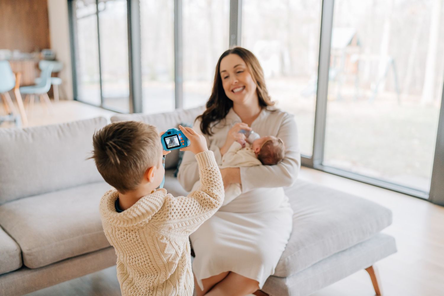 A mother sits on a couch holding her newborn while her young son photographs her with a toy camera.