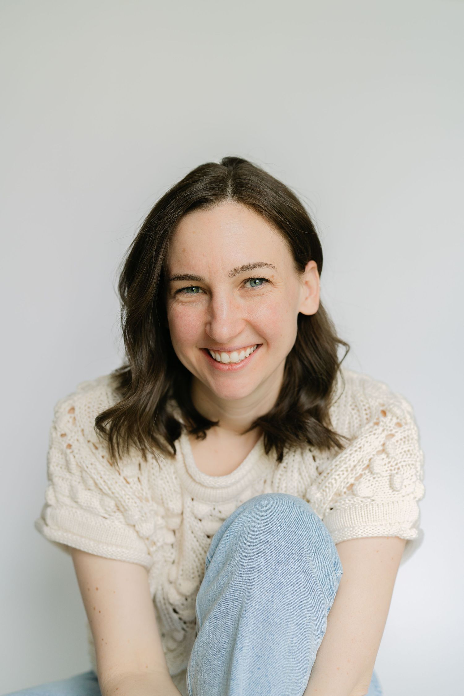 A smiling brunette woman in a cream knit sweater and jeans sits casually against a white wall.