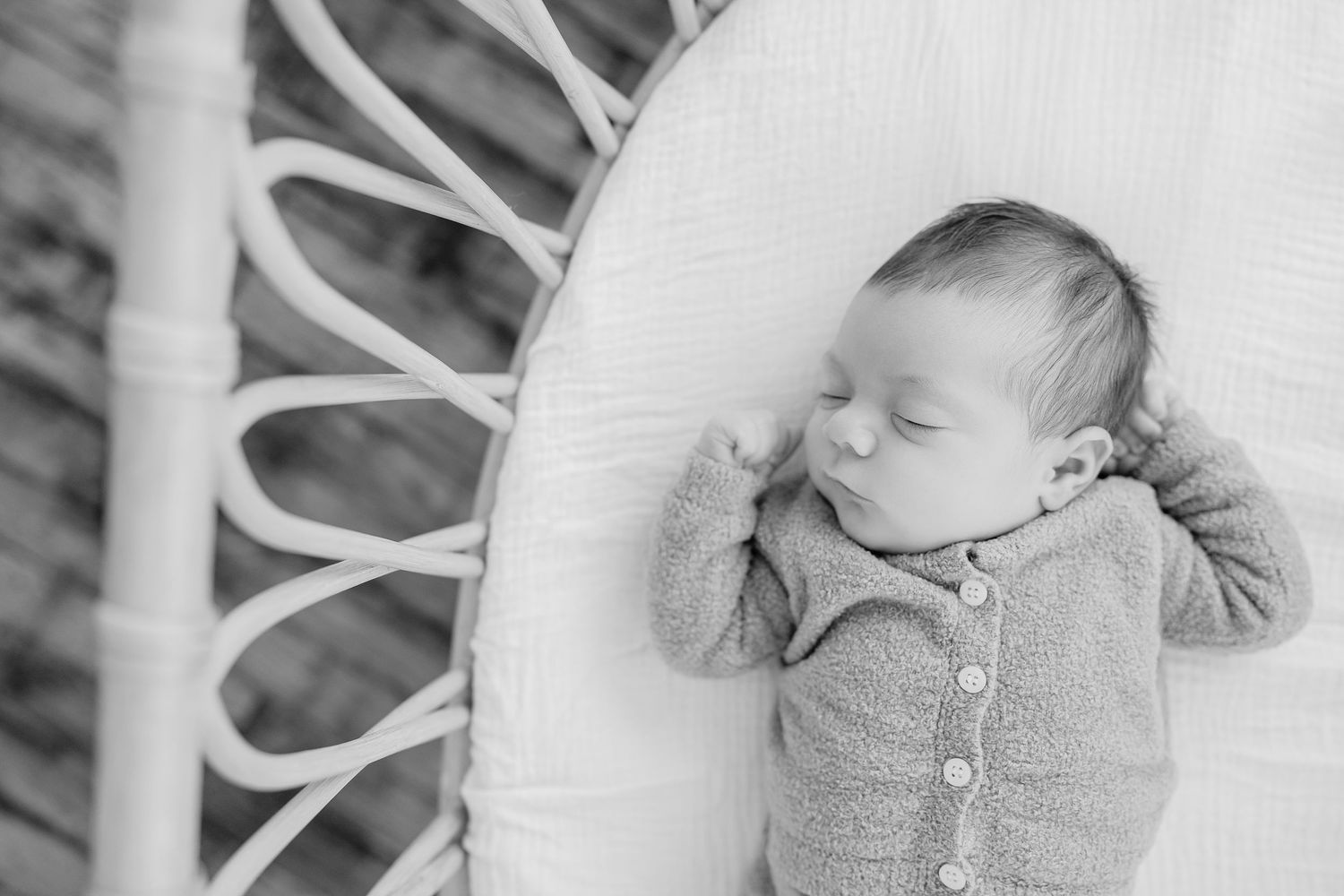 Peaceful newborn baby sleeping in a white wicker bassinet wearing a cozy knit onesie, black and white photo.