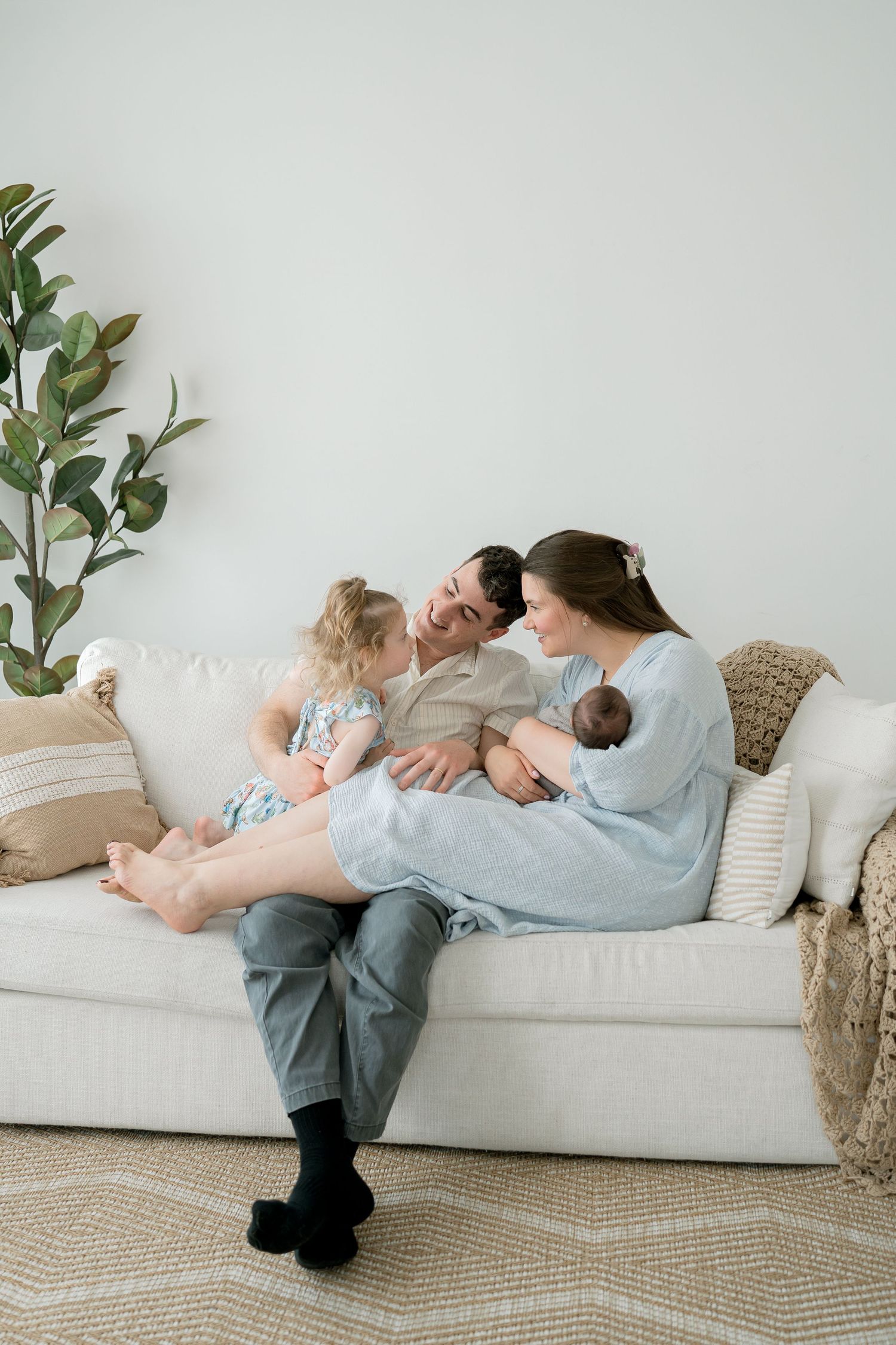 Happy family with toddler and newborn baby sitting together on a white couch in a bright living room.