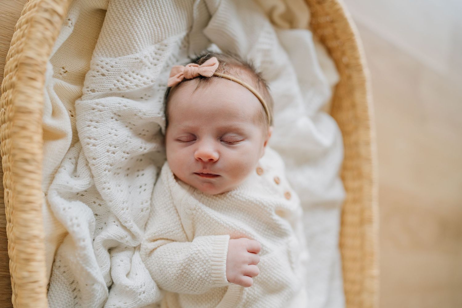 Peaceful newborn baby sleeping in a wicker bassinet wrapped in a soft white knit blanket with a bow headband.