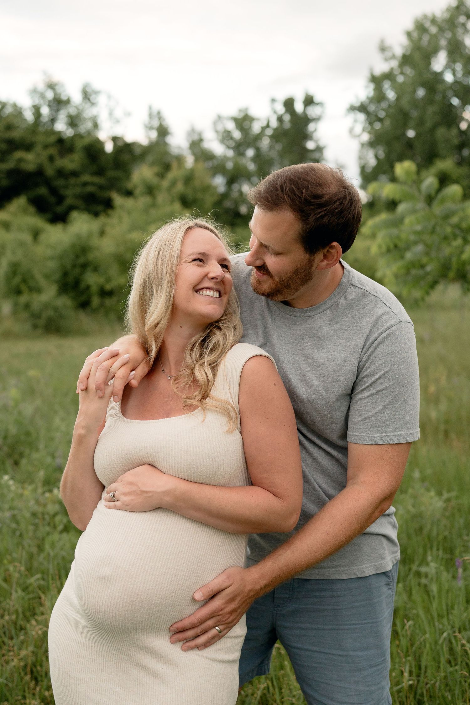 A smiling pregnant woman in a white dress embraces her partner outdoors in a lush green field during a maternity photoshoot.