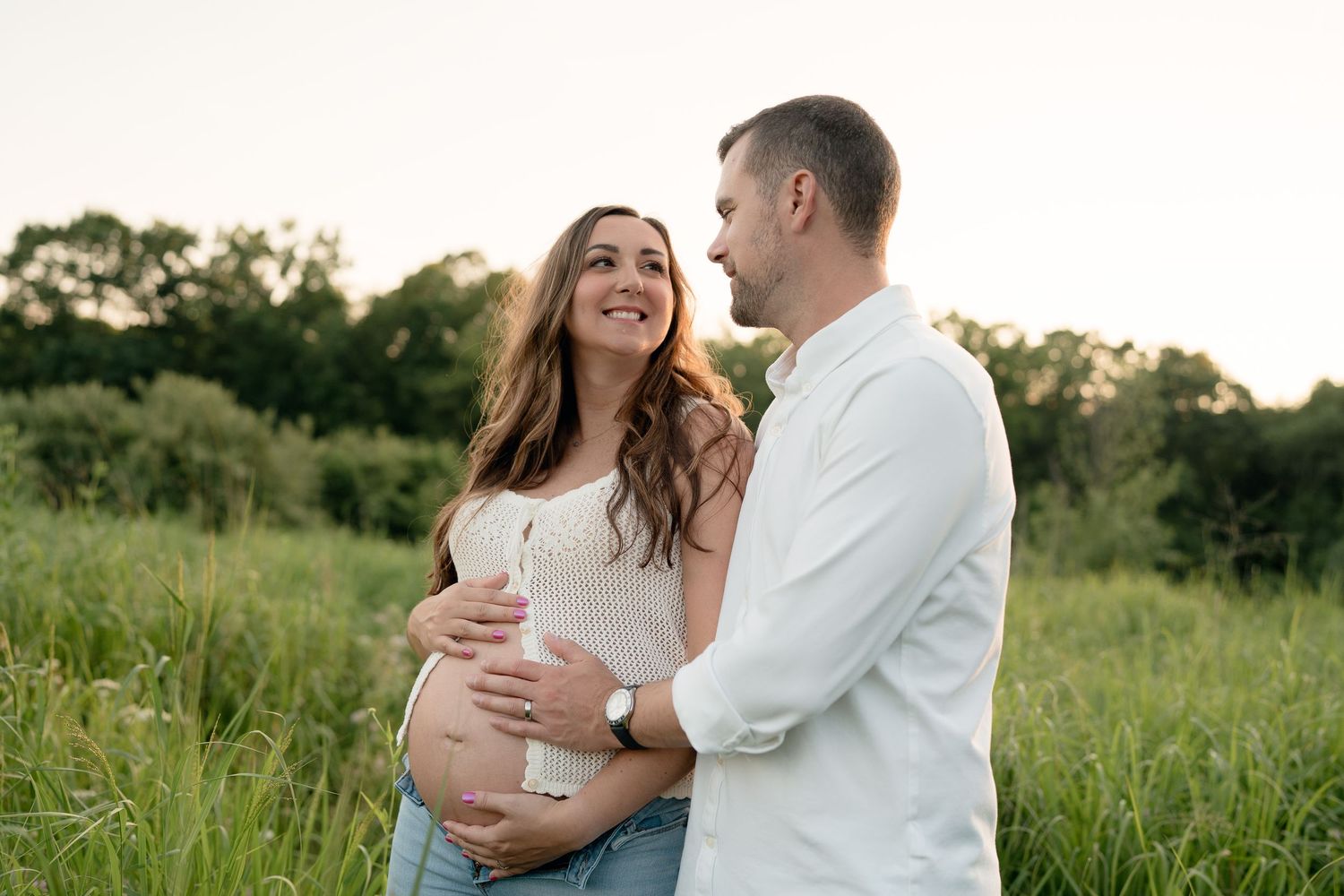 Couple embracing in green field at sunset, man holds pregnant womans belly.