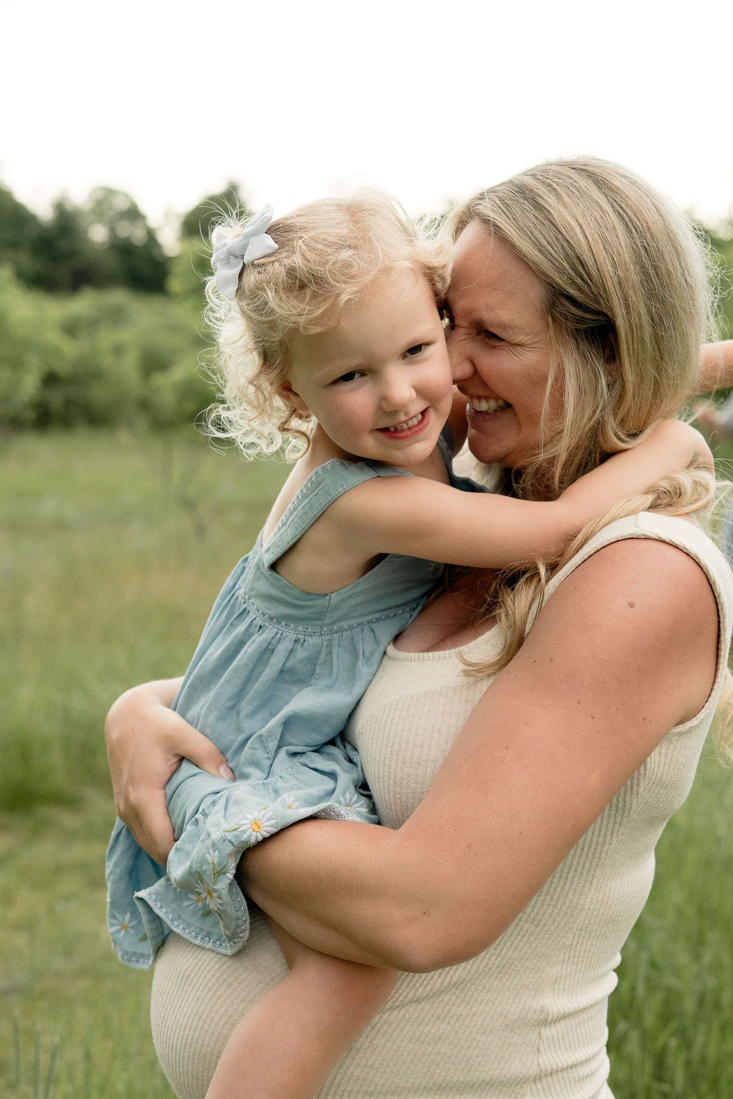 Pregnant mother holding smiling blonde toddler girl in blue dress outdoors in green field.