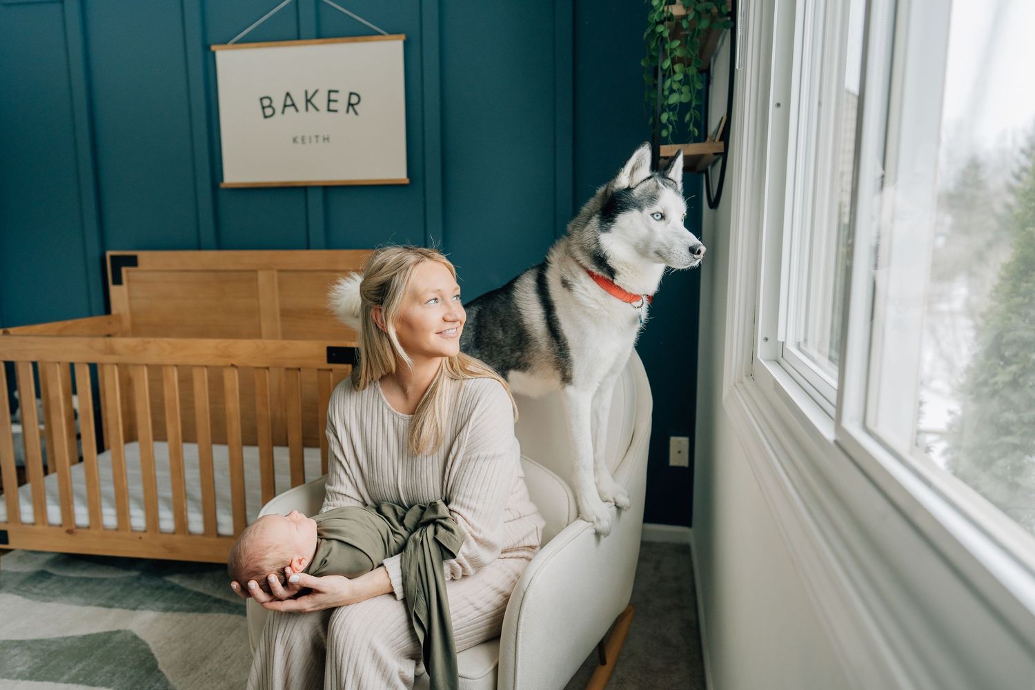 Blonde woman in grey robe sits with Husky dog in teal nursery room with wooden crib.