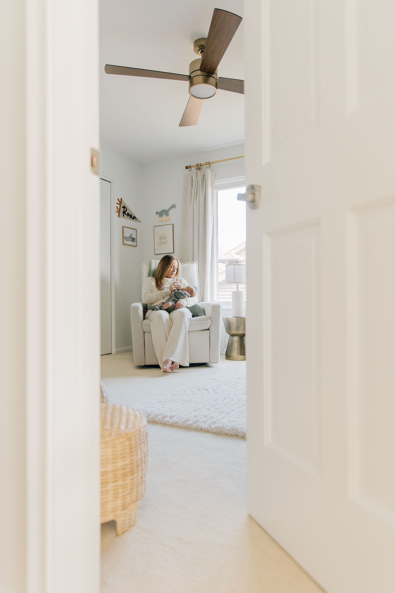 Mother sitting in a nursery glider chair holding her newborn baby viewed through an open doorway.