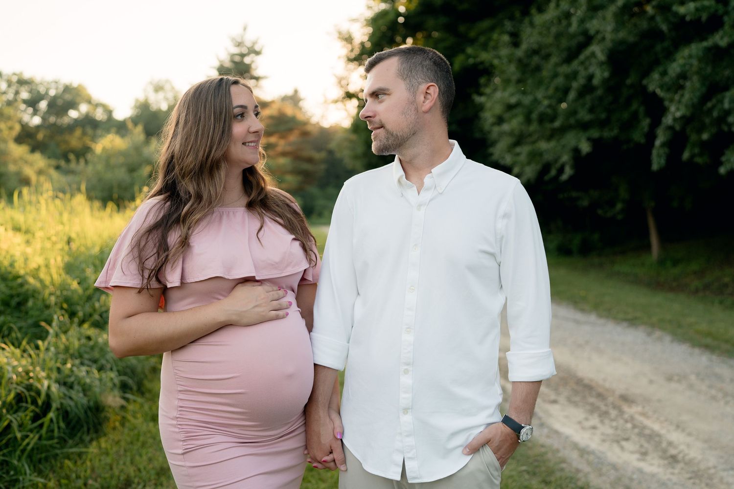 Expecting couple holds hands and gazes at each other on a sunlit garden path.