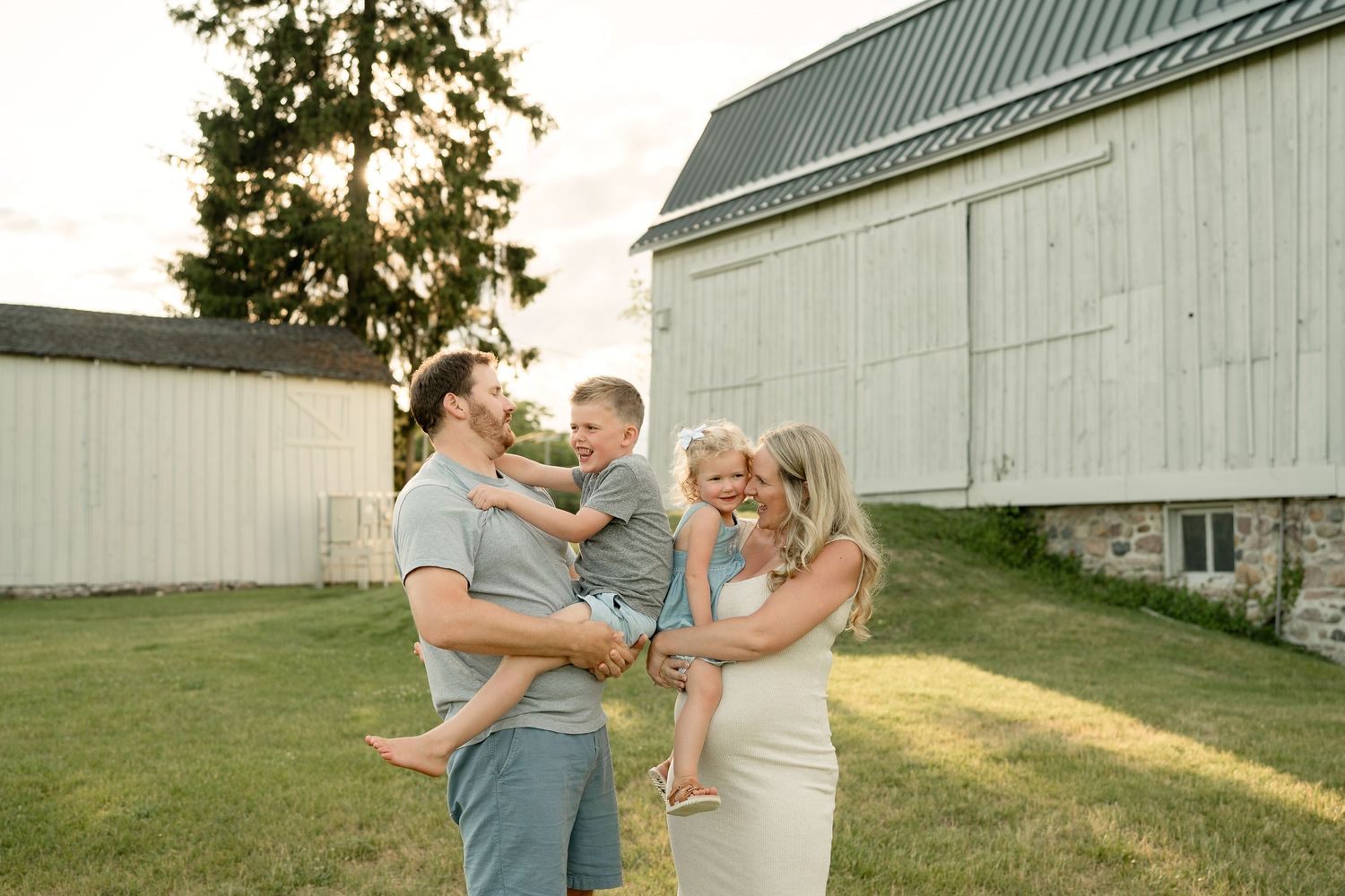 Family of four laughing outdoors near a white barn at golden hour, parents holding young children on a green lawn.